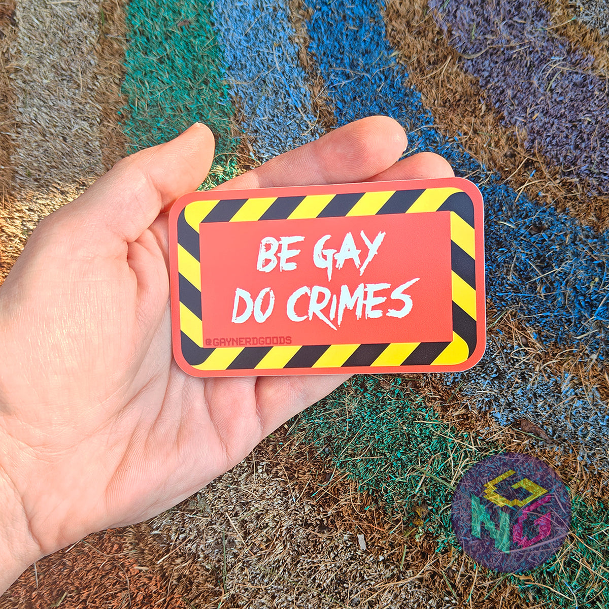 Rectangular red sticker featuring white text reading "BE GAY DO CRIMES" surrounded by black and yellow striped border. The sticker being held in an open palm in front of a rough rainbow background
