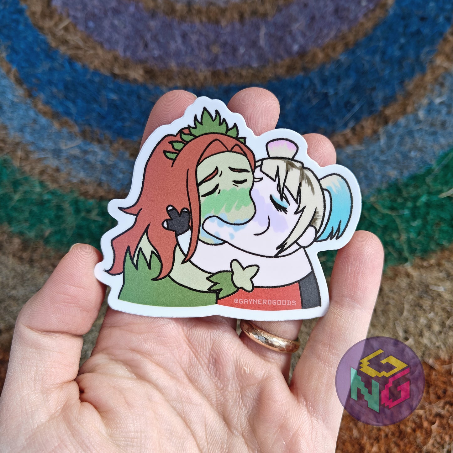 harlivy harley and ivy kissing sticker in a hand in front of a rainbow and neutral background