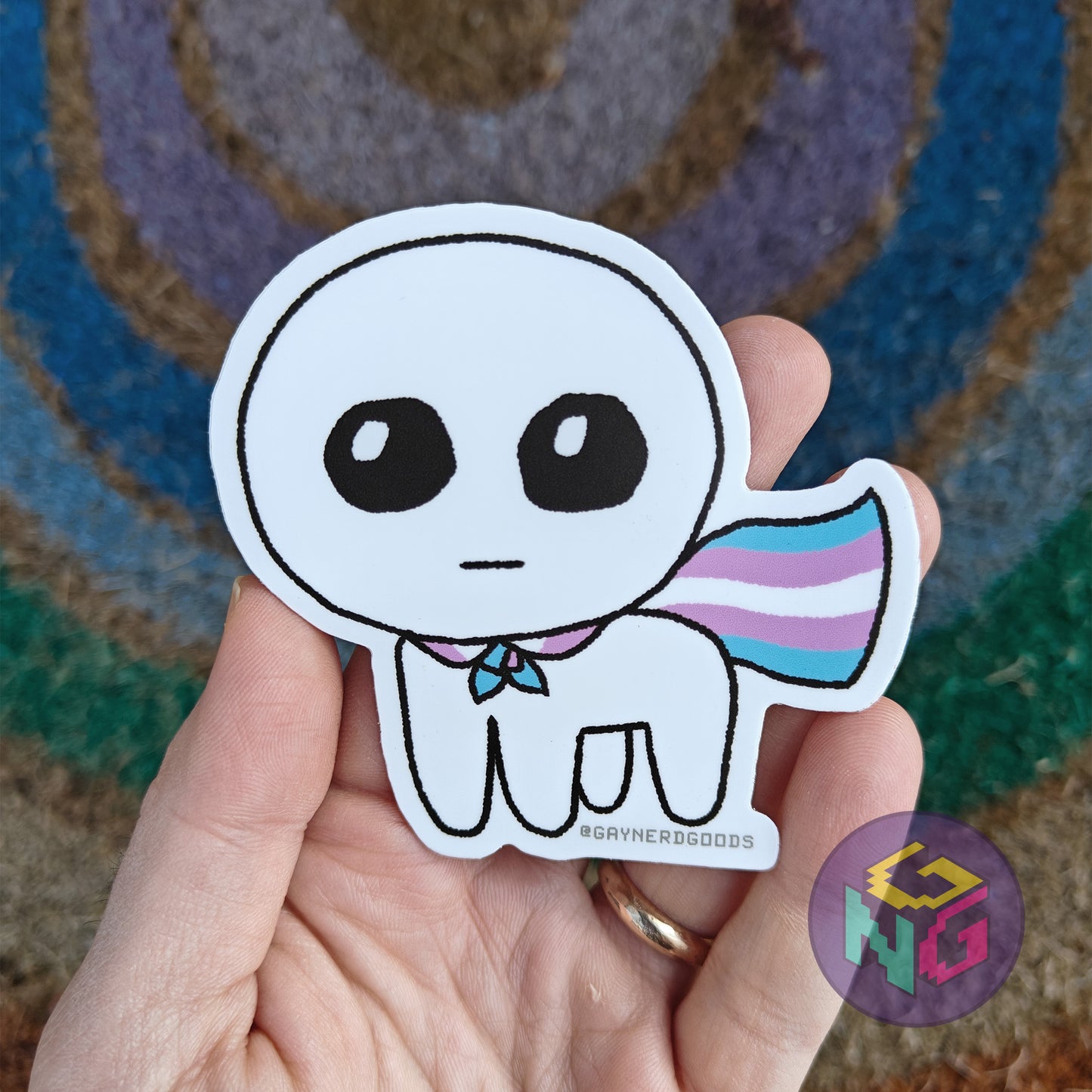 transgender autism creature sticker being held in front of a neutral and rainbow background