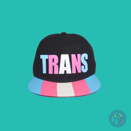 Black flat bill snapback hat. The brim has the transgender pride flag on both sides and the front of the hat has the word “TRANS” in blue, pink, and white letters. Front view