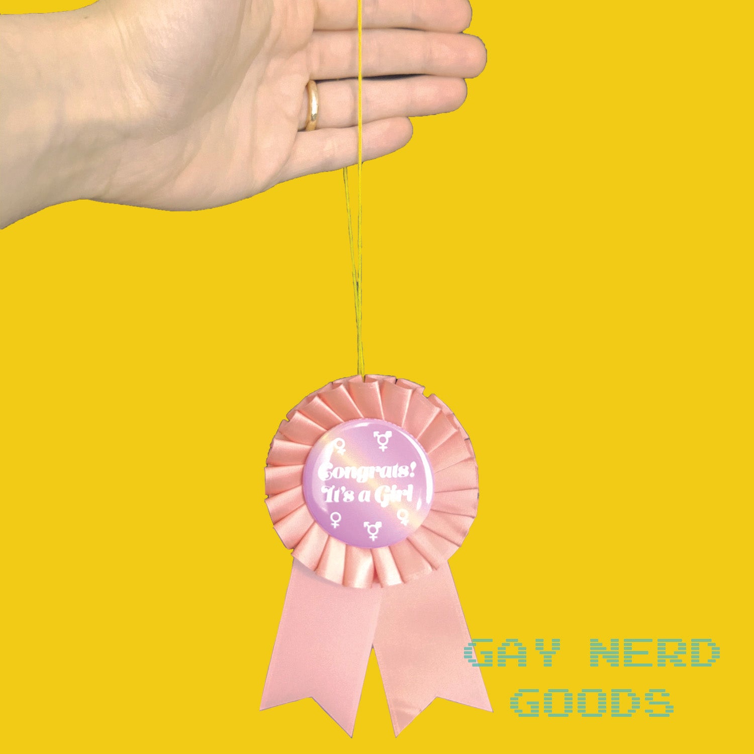 hand holding the thread loop of the "congrats it's a girl" award ribbon on a yellow background