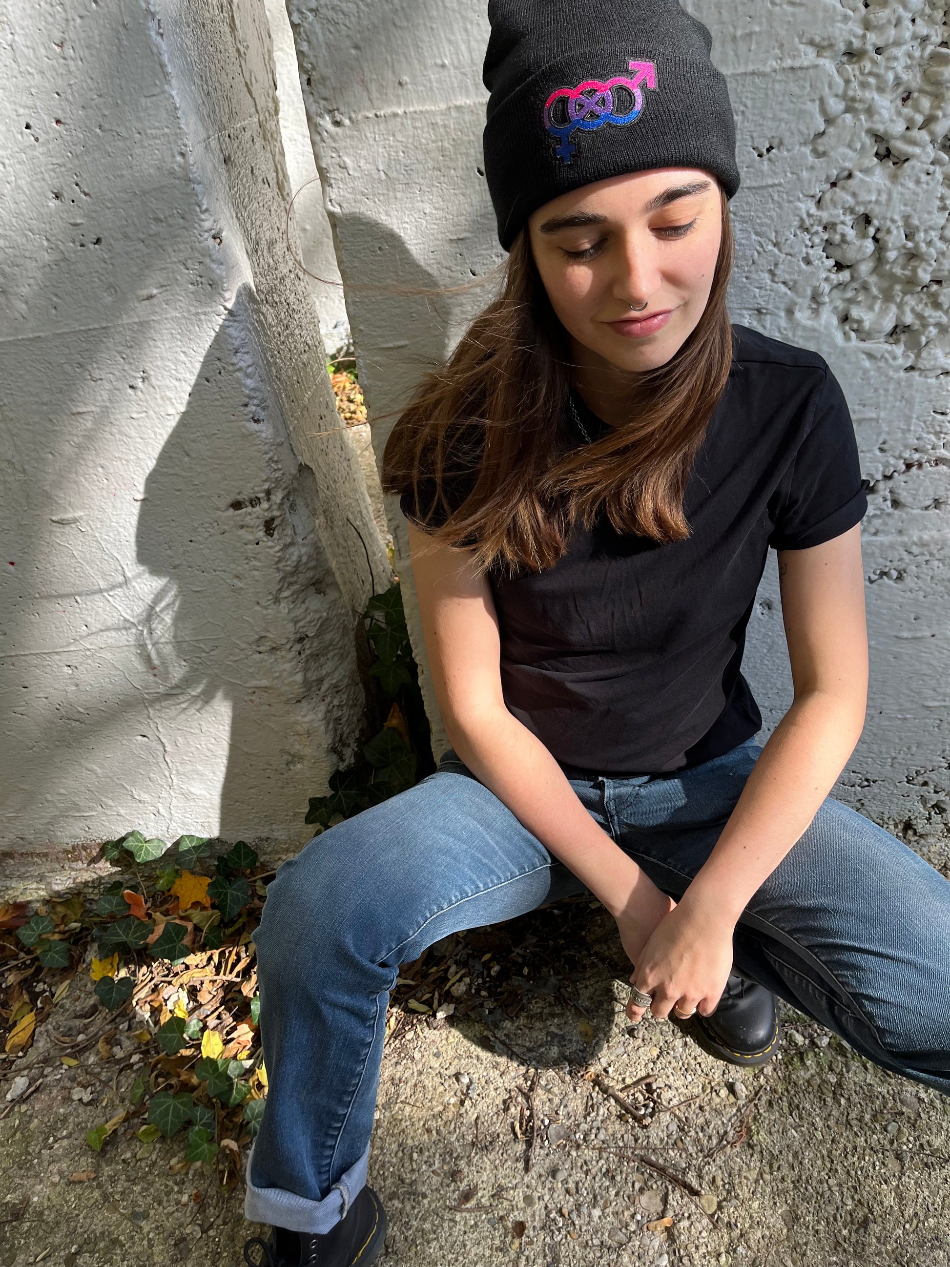 person crouching against a white brick concrete wall wearing the bisexual pride beanie