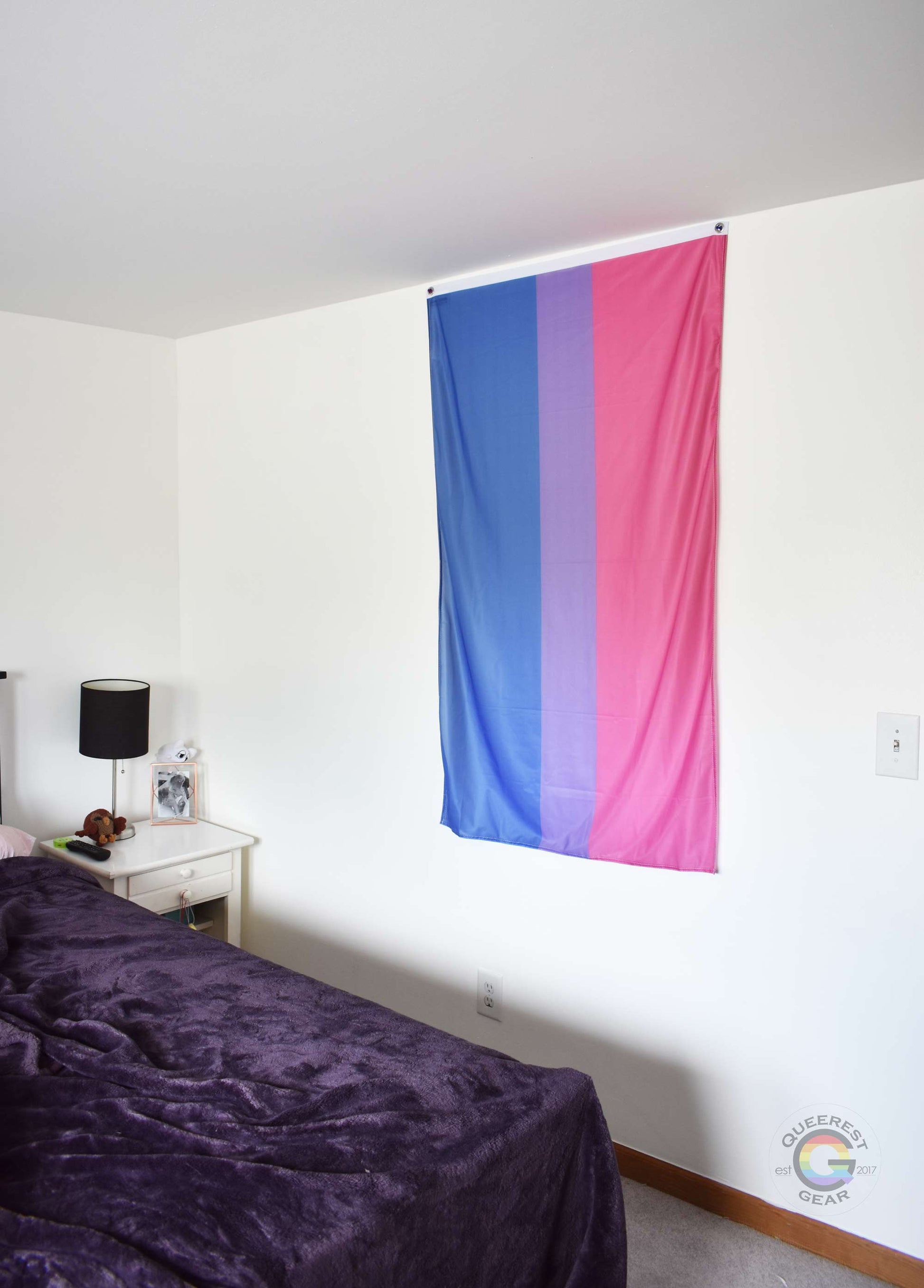  3’x5’ bisexual pride flag hanging vertically on the wall of a bedroom with a nightstand and a bed