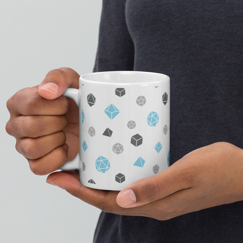 white mug being held by the handle by a dark-skinned set of hands. It has an all-over print of polyhedral d&d dice in the demiboy colors of dark grey, light grey, and blue