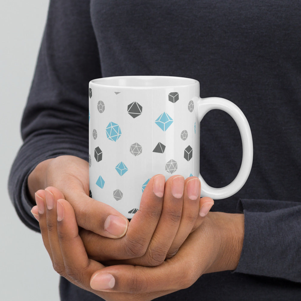 white mug being cupped by a dark-skinned set of hands. It has an all-over print of polyhedral d&d dice in the demiboy colors of dark grey, light grey, and blue