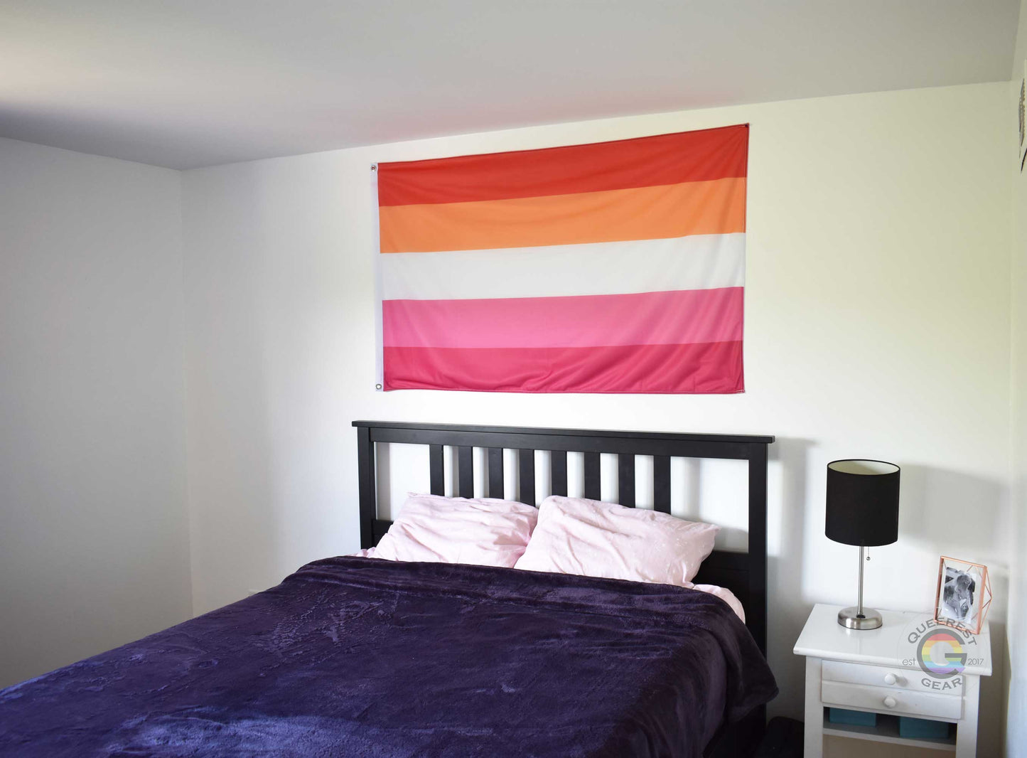 3’x5’ lesbian pride flag hanging horizontally on the wall of a bedroom centered above a bed with a purple blanket