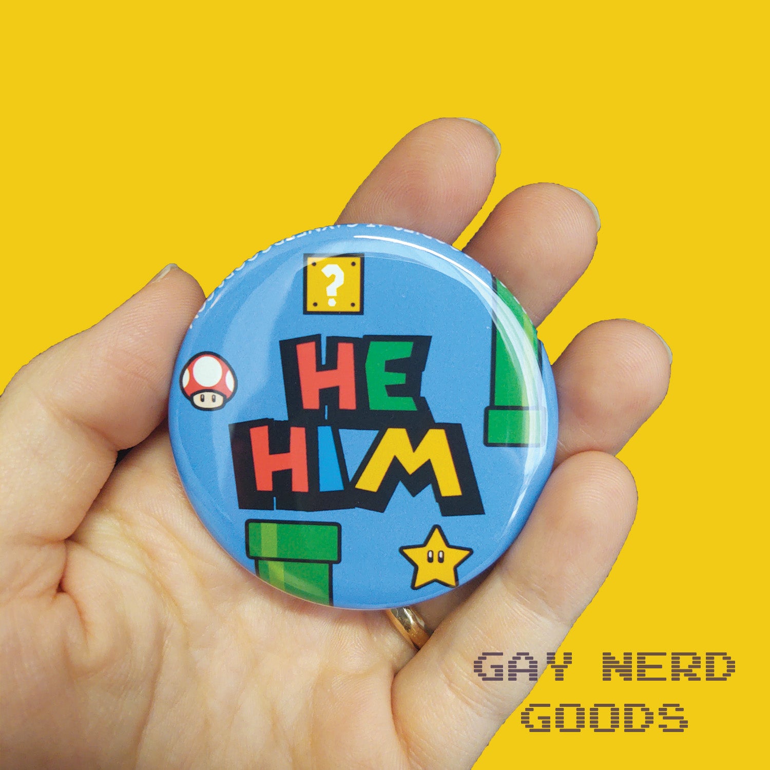 he him mario pronoun button with blue background, green pipes, question block, star, and mushroom held in a hand