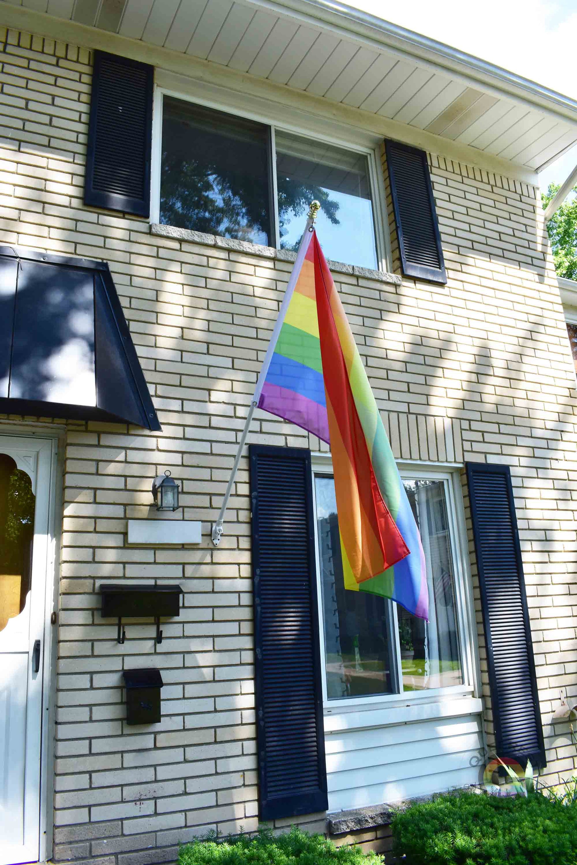 3’x5’ rainbow pride flag hanging from a flagpole on the outside of a light brick house with dark shutters
