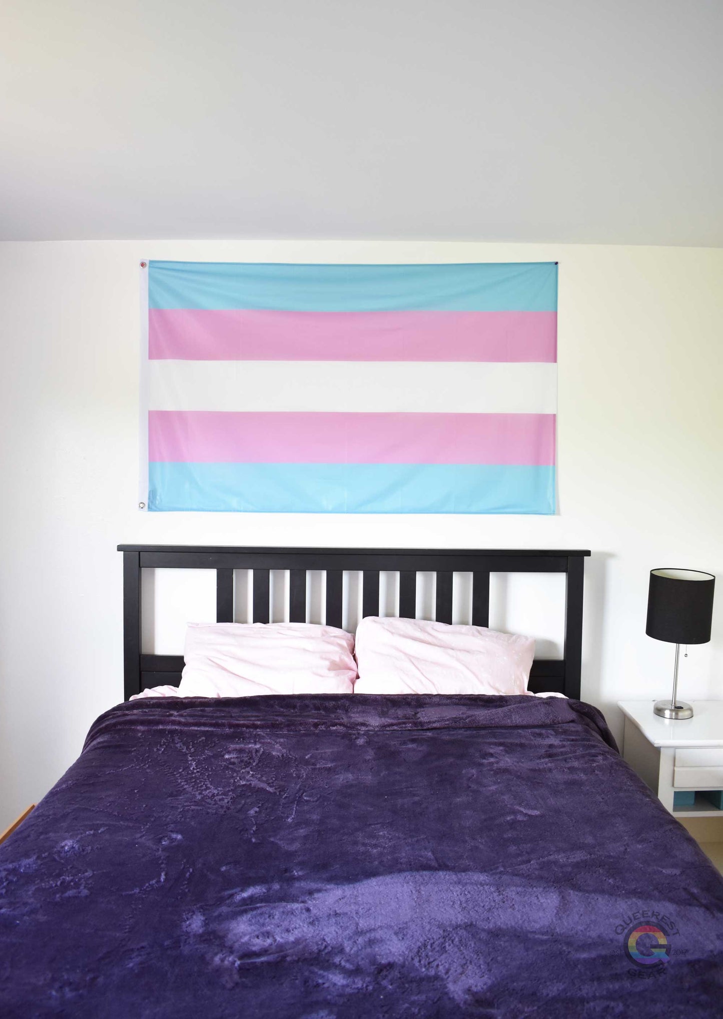 3’x5’ transgender pride flag hanging horizontally on the wall of a bedroom centered above a bed with a purple blanket