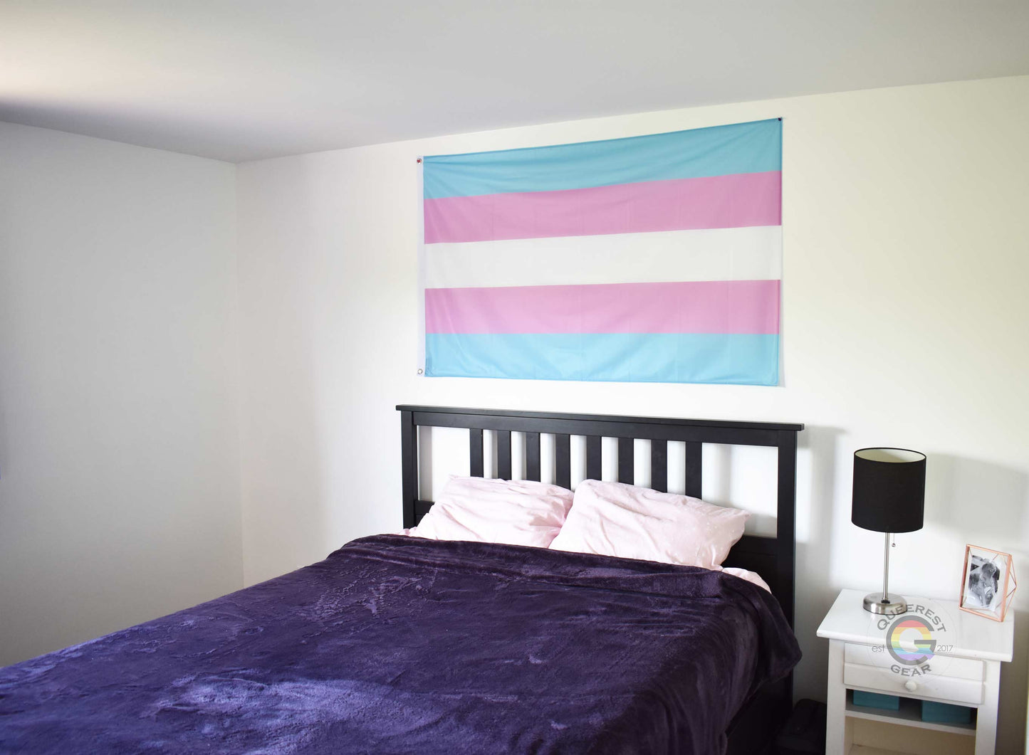 3’x5’ transgender pride flag hanging horizontally on the wall of a bedroom centered above a bed with a purple blanket