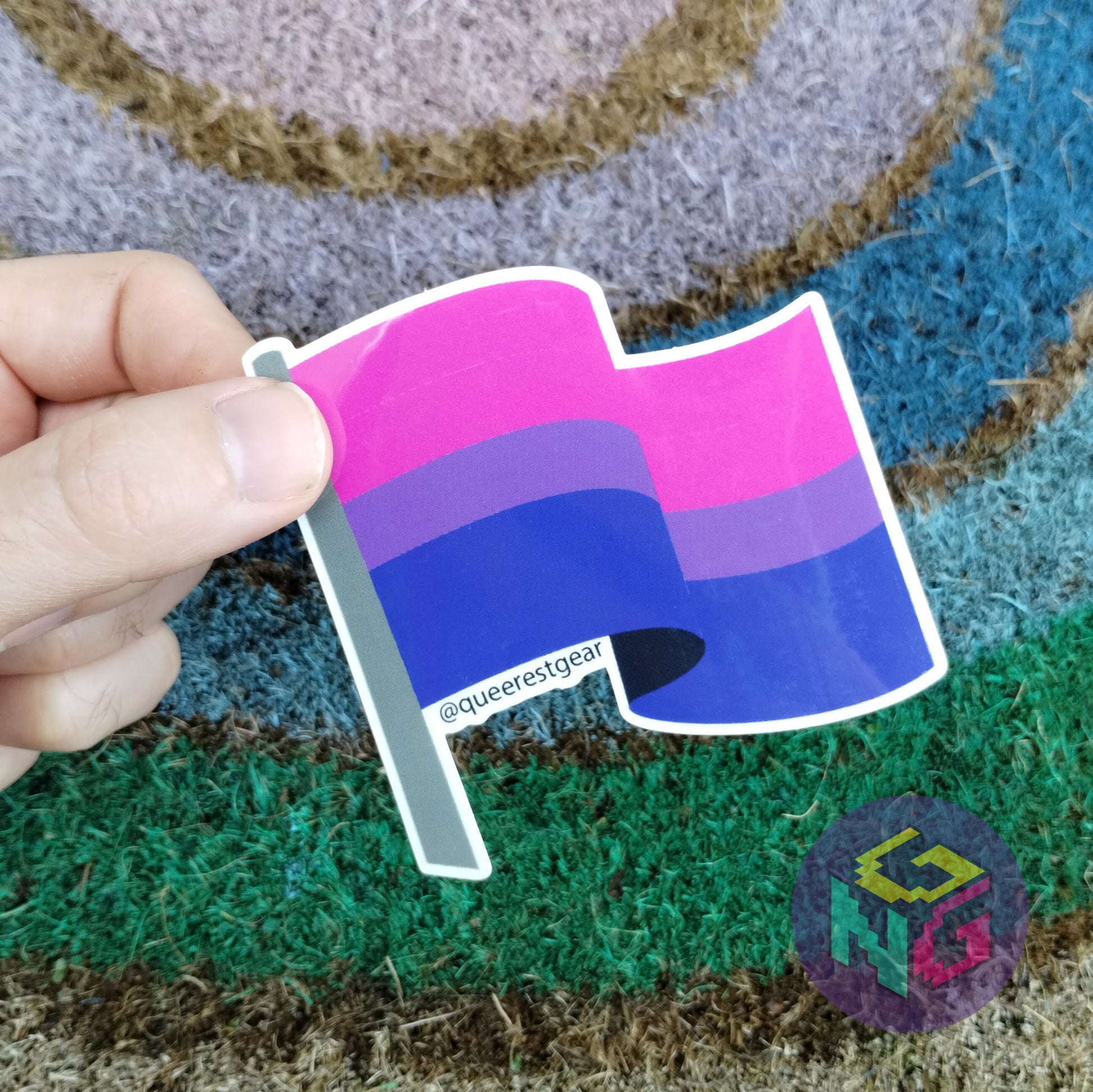 bisexual flag sticker held between thumb and finger against rainbow welcome mat