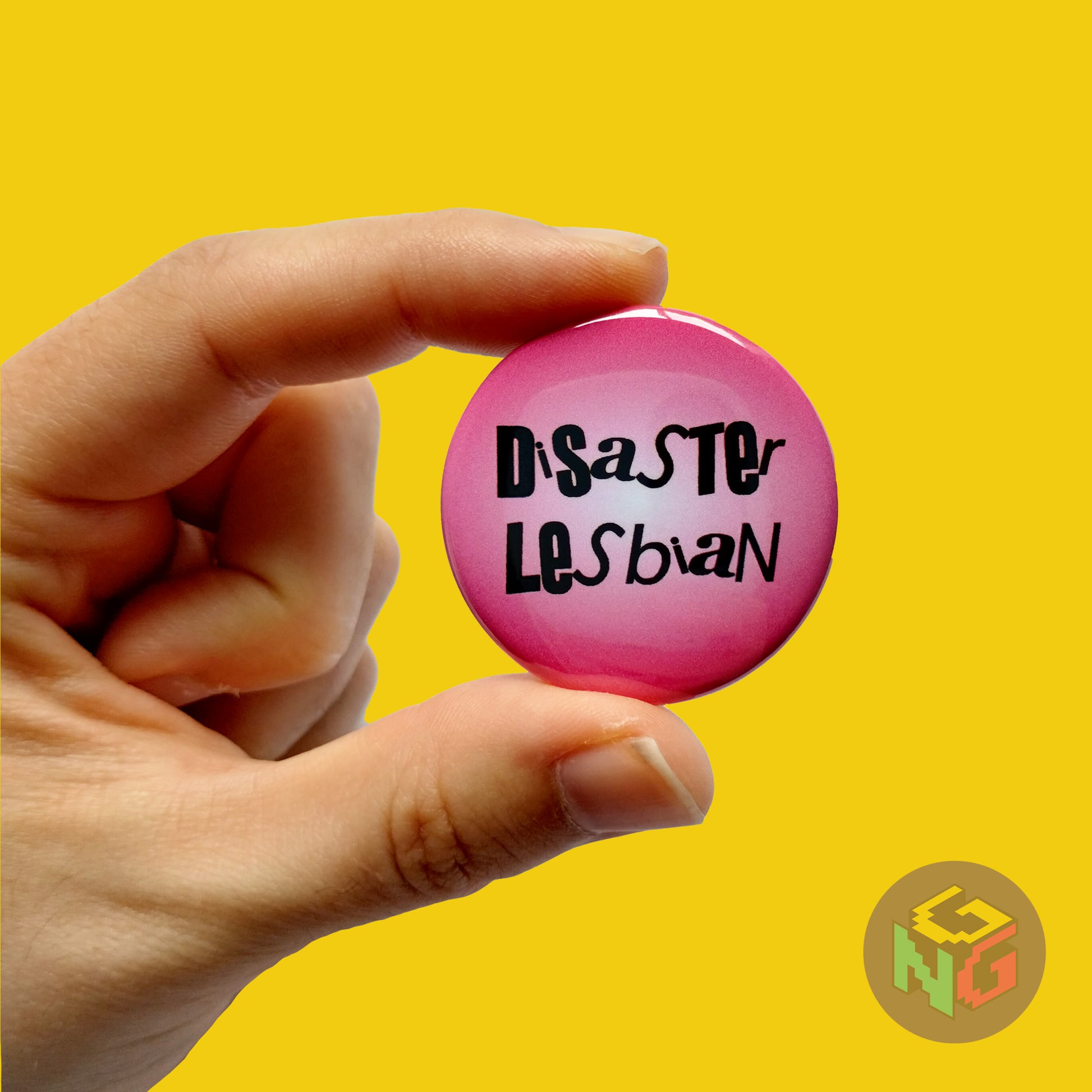pink disaster lesbian pin held in a hand in front of yellow background