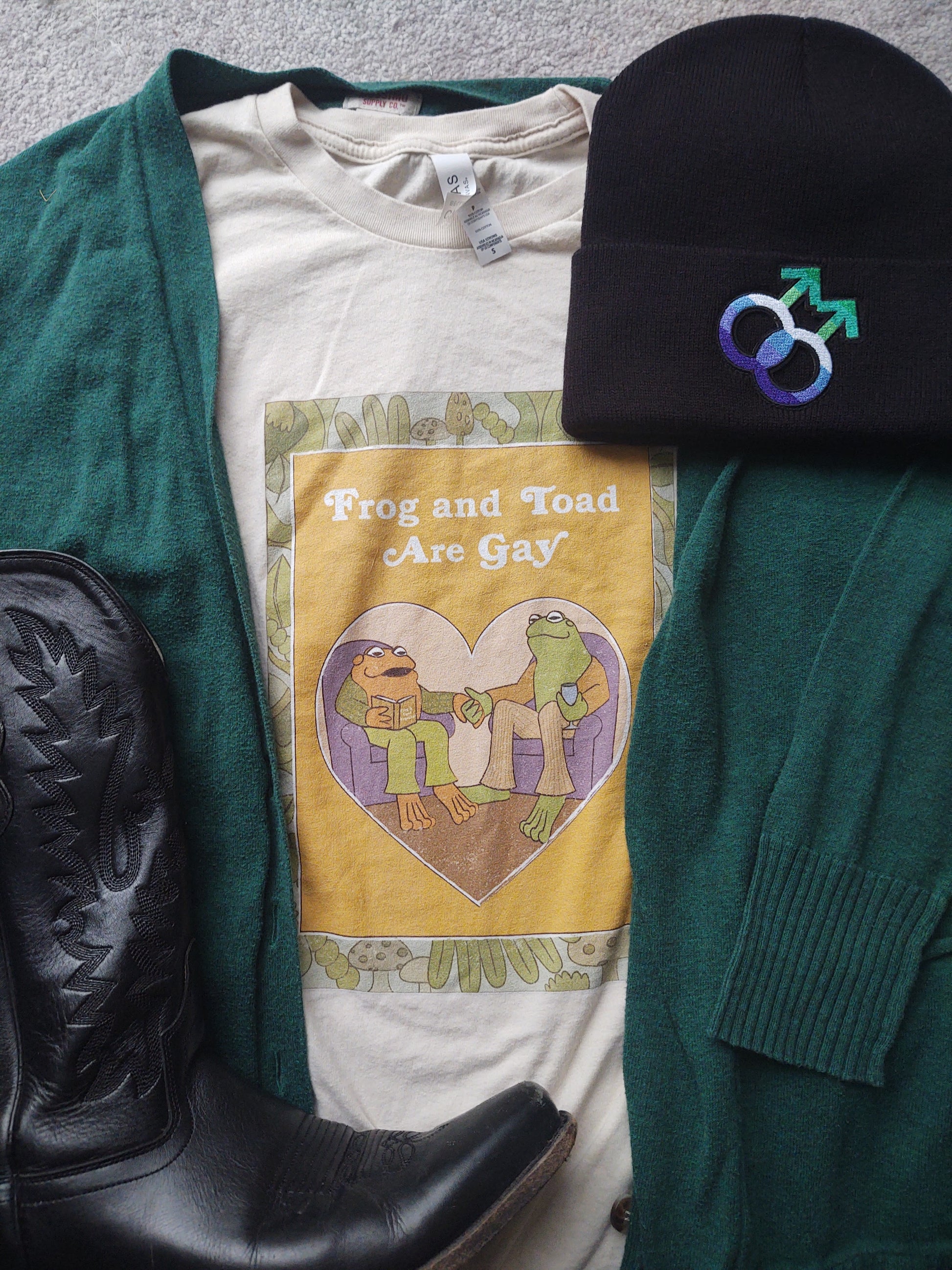 frog and toad shirt flat lay with green cardigan, black boot, and gay mlm beanie