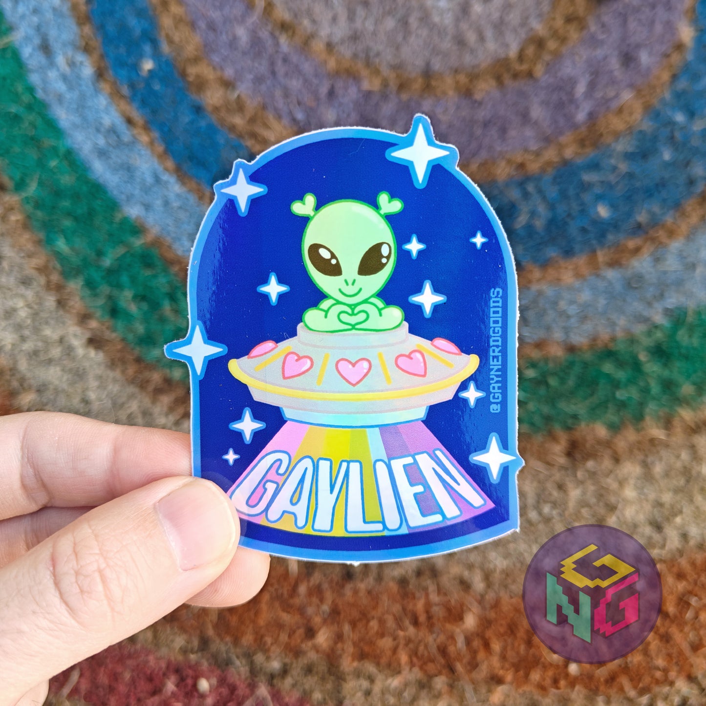 gaylien holographic alien sticker with a littel green alien in a spaceship with a rainbow beam held in front of a rainbow welcome mat