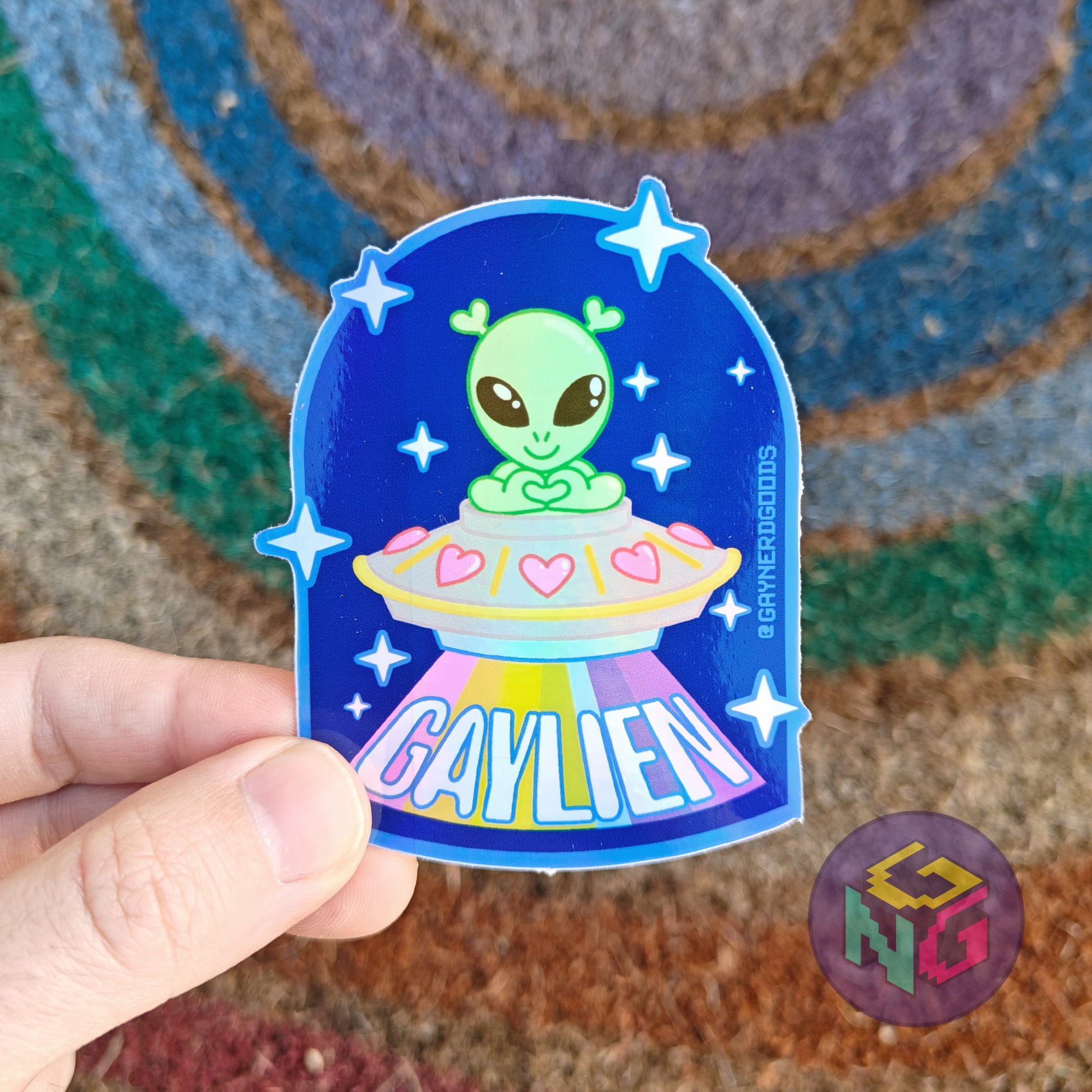 gaylien holographic alien sticker with a littel green alien in a spaceship with a rainbow beam held in front of a rainbow welcome mat