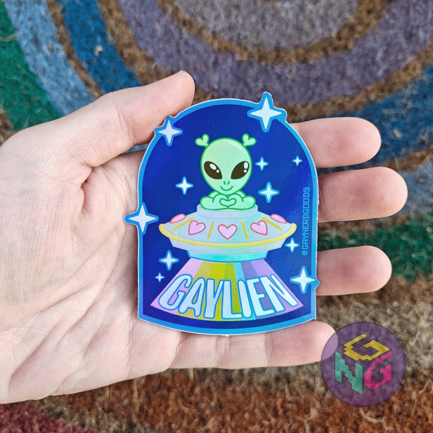 gaylien holographic alien sticker with a littel green alien in a spaceship with a rainbow beam held in front of a rainbow welcome mat