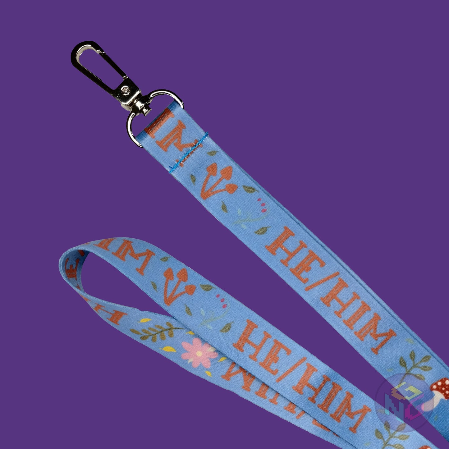 close up detail of the blue he him nature lanyard showing the lobster clasp, brown mushrooms, green leaves, and all caps text