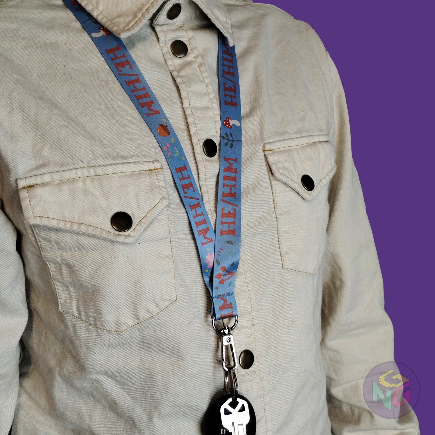 blue he him mushroom lanyard being worn by a flat chested person wearing a cream button up. The end of the lanyard falls between their sternum and bellybutton and has a key fob clipped to the end