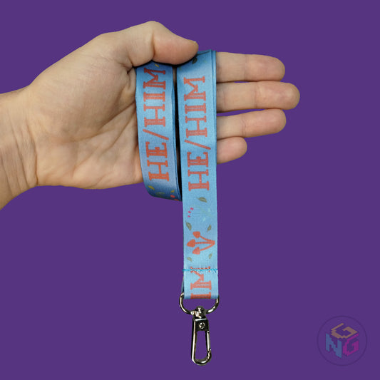 blue cottagecore he him pronoun lanyard wrapped around a hand with the lobster clasp dangling against a purple background