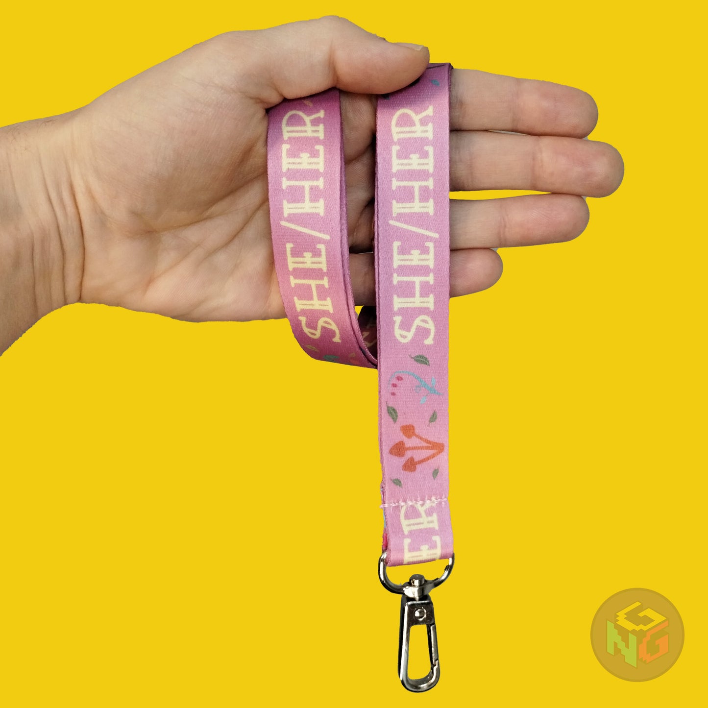 pink cottagecore she her pronoun lanyard wrapped around a hand with the lobster clasp dangling against a yellow background