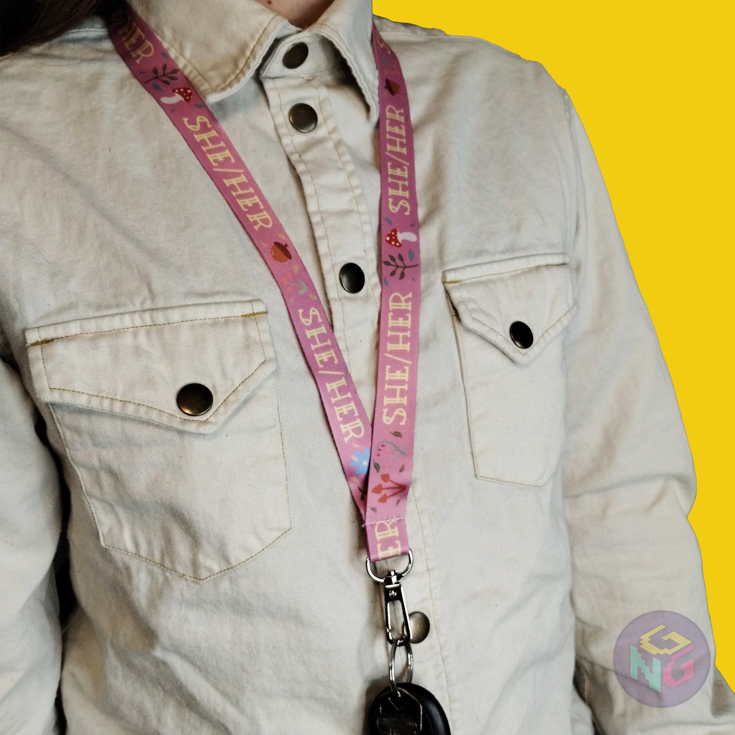 pink she her mushroom lanyard being worn by a flat chested person wearing a cream button up. The end of the lanyard falls between their sternum and bellybutton and has a key fob clipped to the end