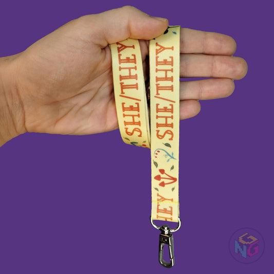 yellow cottagecore she they pronoun lanyard wrapped around a hand with the lobster clasp dangling against a purple background