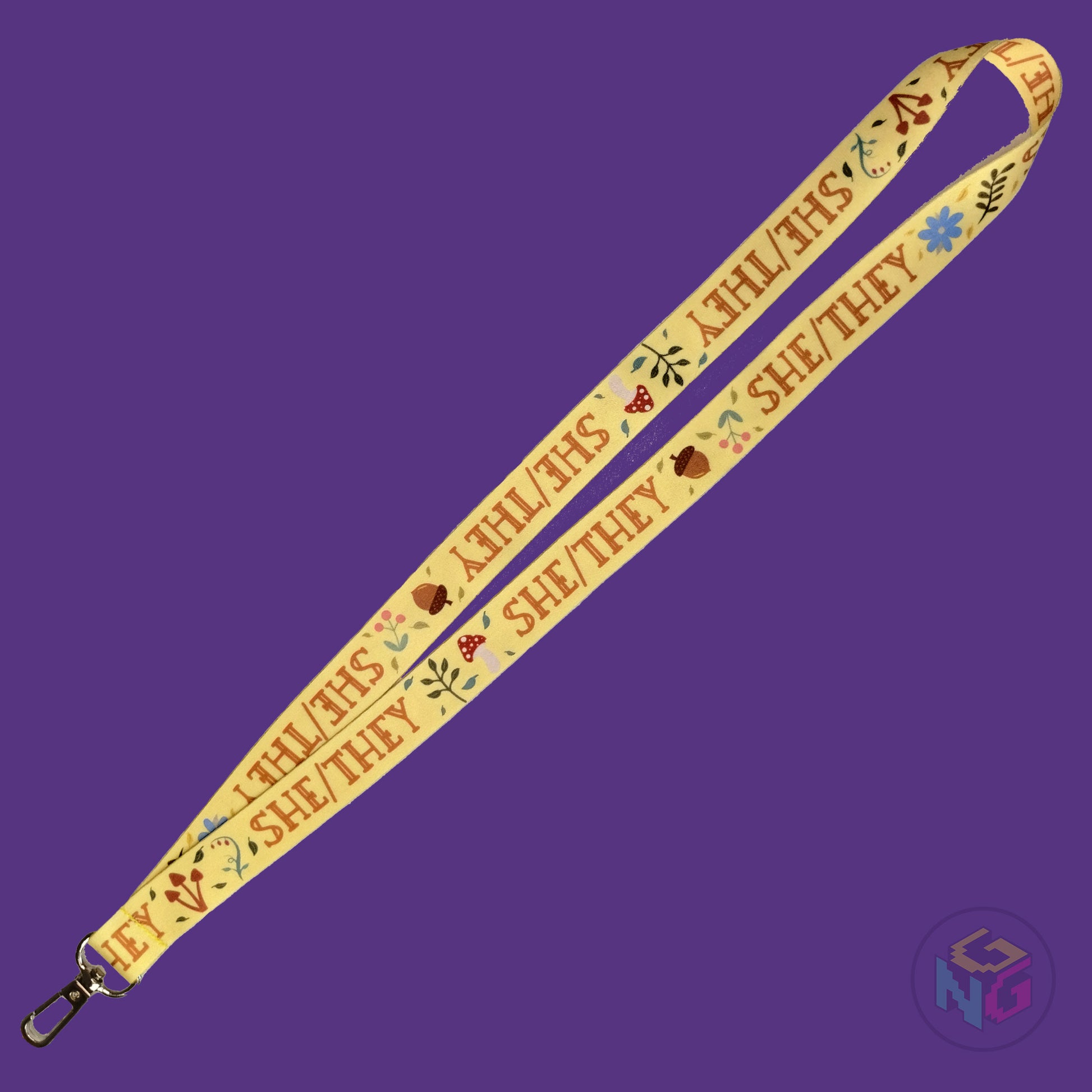 the yellow she they cottagecore lanyard lying flat showing the complete design and repeating pattern of acorns, berries, mushrooms, leaves, and she they pronouns