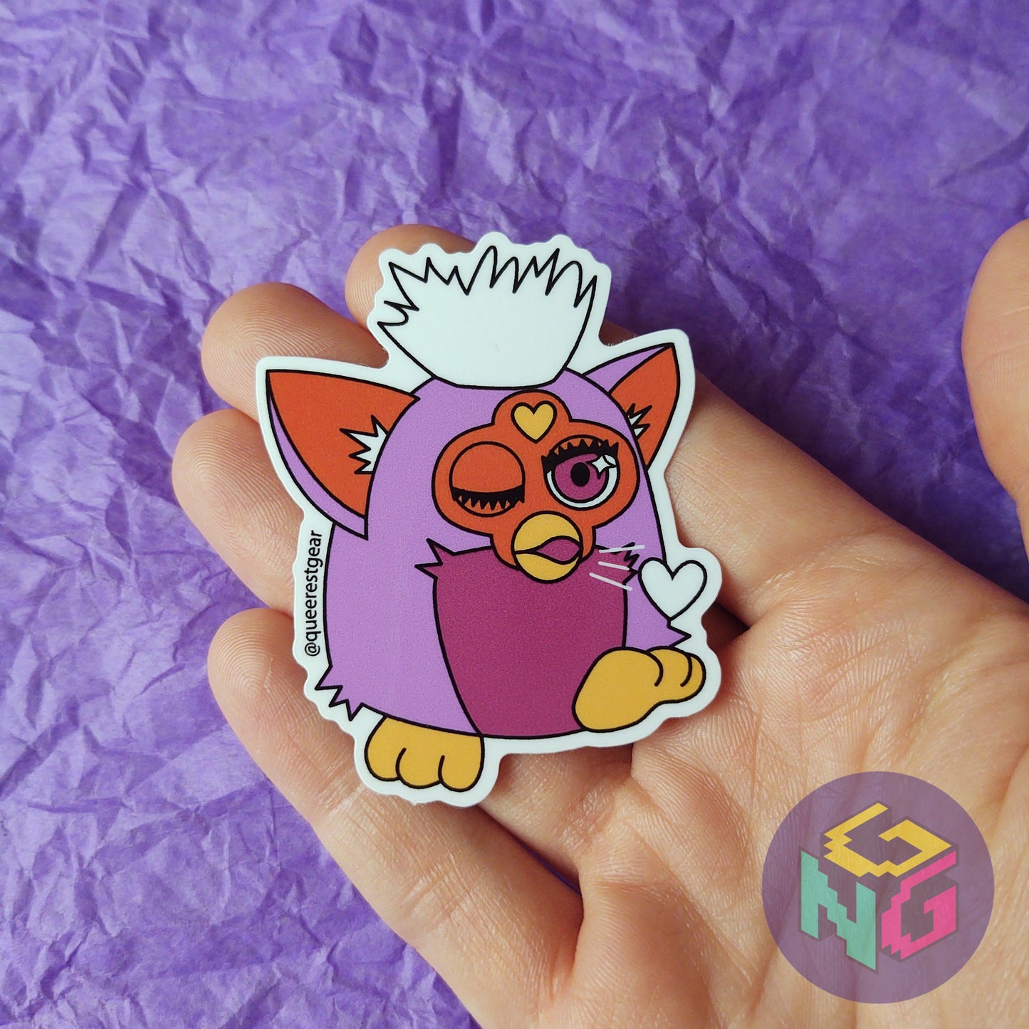 lesbian furby sticker blowing a kiss being held in a hand in front of a purple background