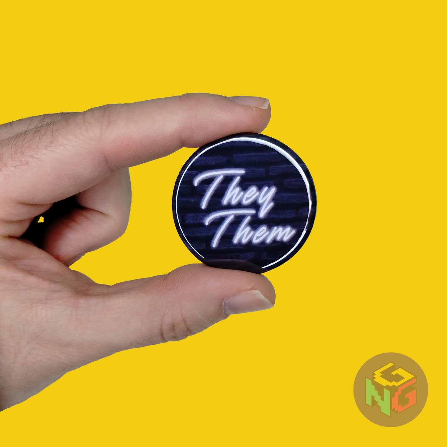round they them pronoun pin featuring white neon text on a dark blue brick background. The pin is held in a hand in front of a yellow background