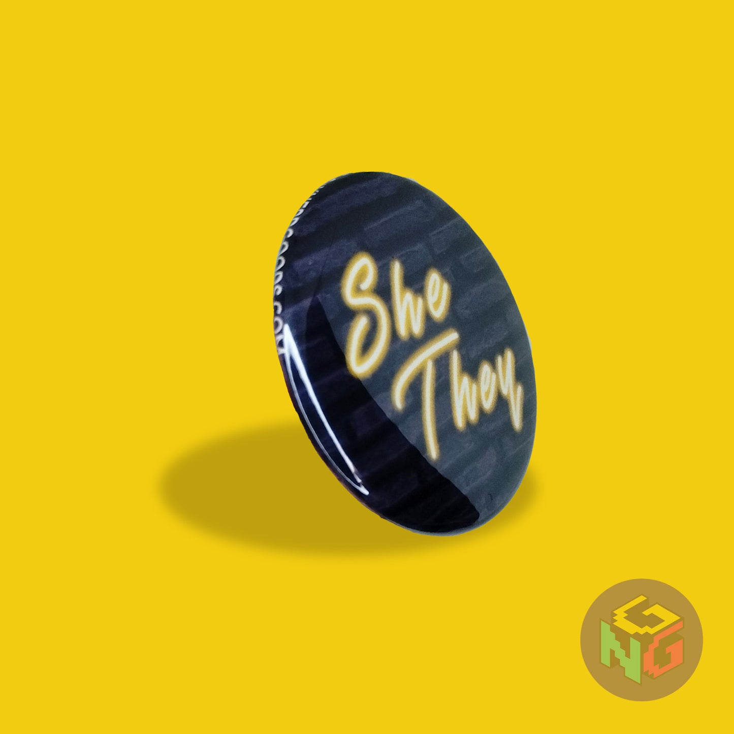 neon she they pride button with yellow text on a dark blue brick background facing to the right on a yellow background