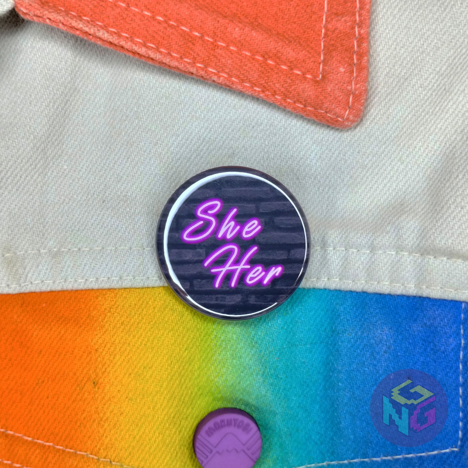 she her neon pronoun pin with pink glowing text on a dark brick. The pin is attached to a white denim jacket with rainbow accents