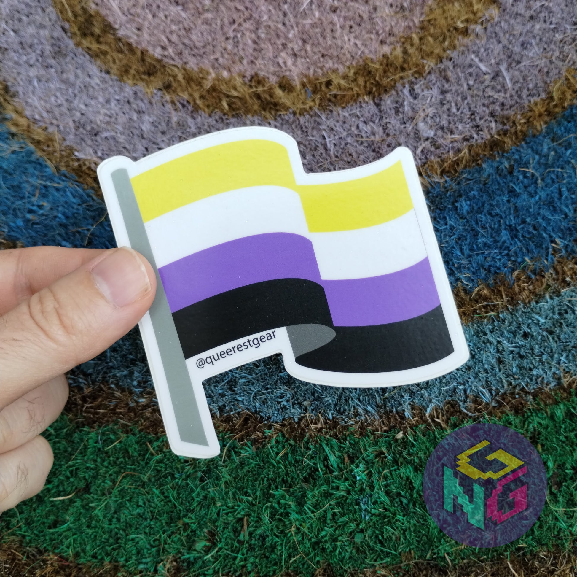 nonbinary flag sticker held between finger and thumb in front of rainbow welcome mat