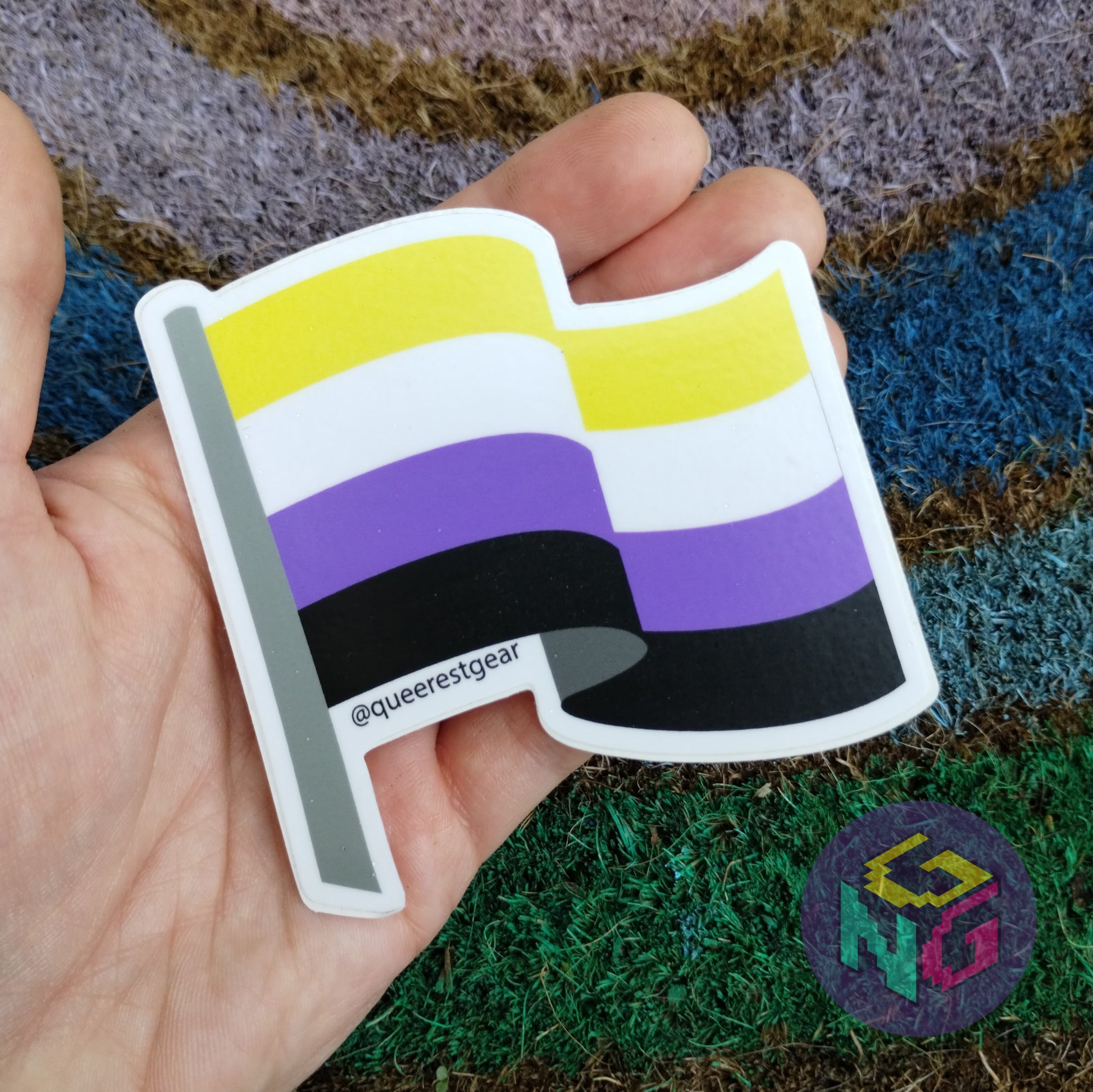 nonbinary flag sticker held in hand in front of rainbow welcome mat