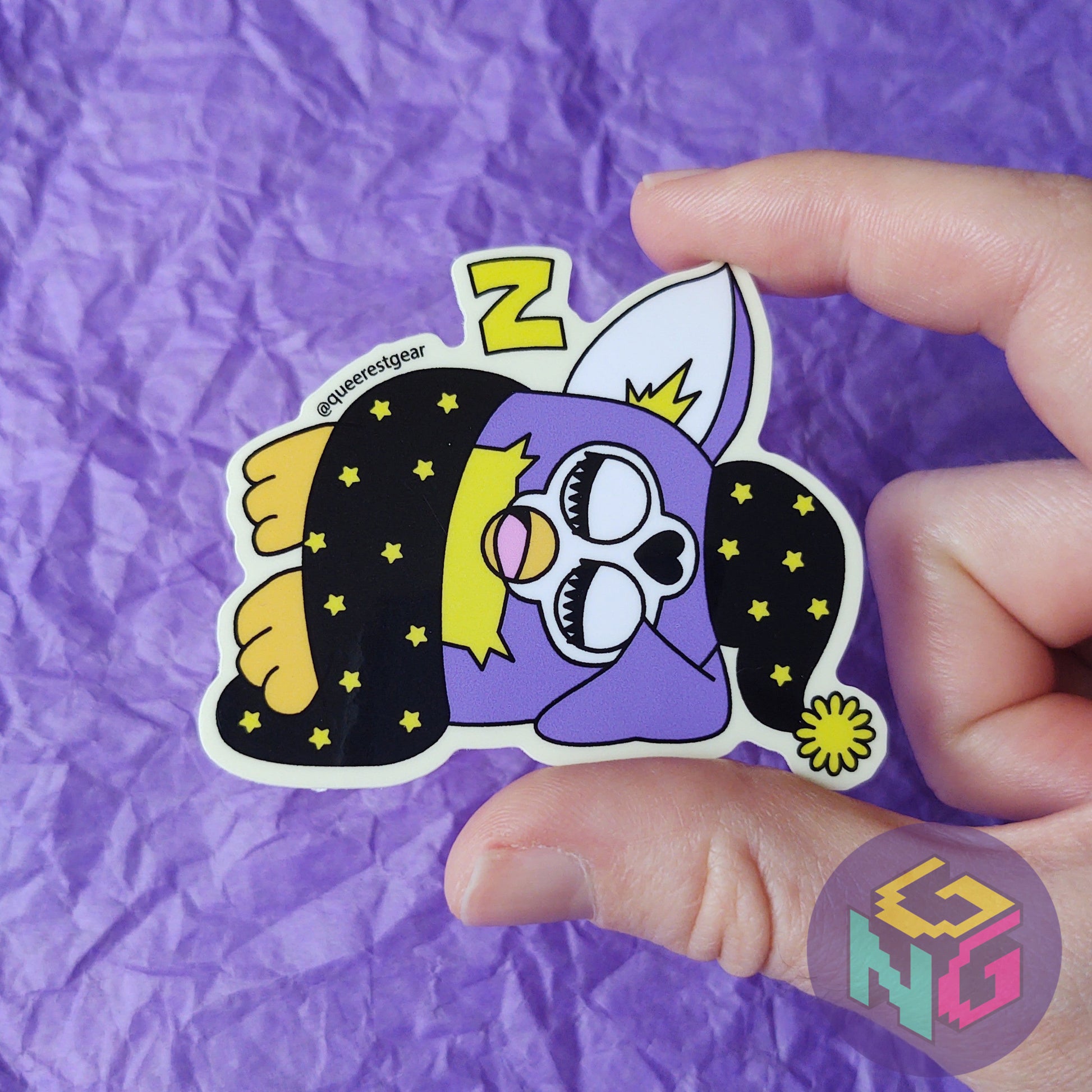 nonbinary furby sticker sleeping being held in a hand in front of a purple background