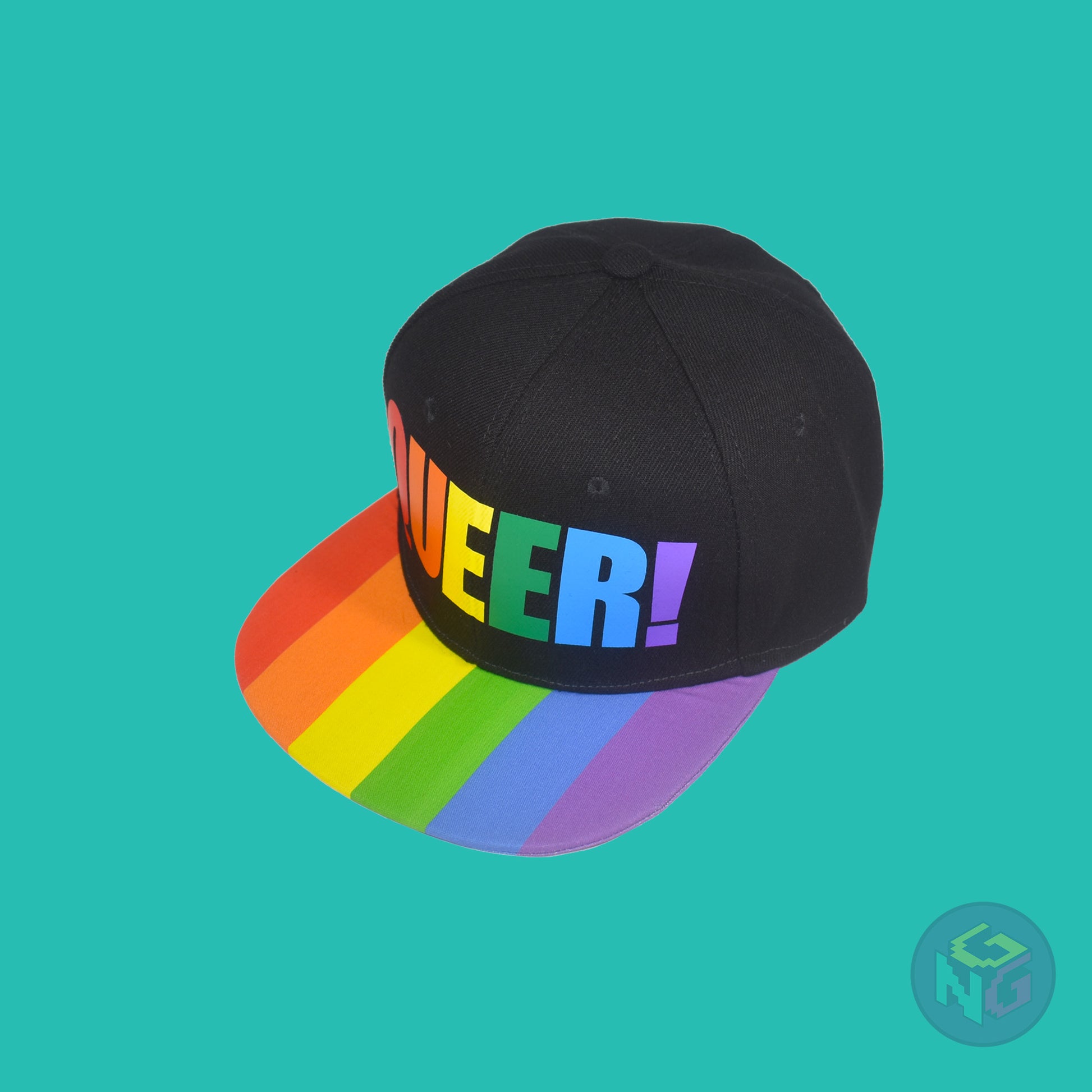 Black flat bill snapback hat. The brim has the rainbow pride flag on both sides and the front of the hat has the word “QUEER!” in rainbow letters. Front left view