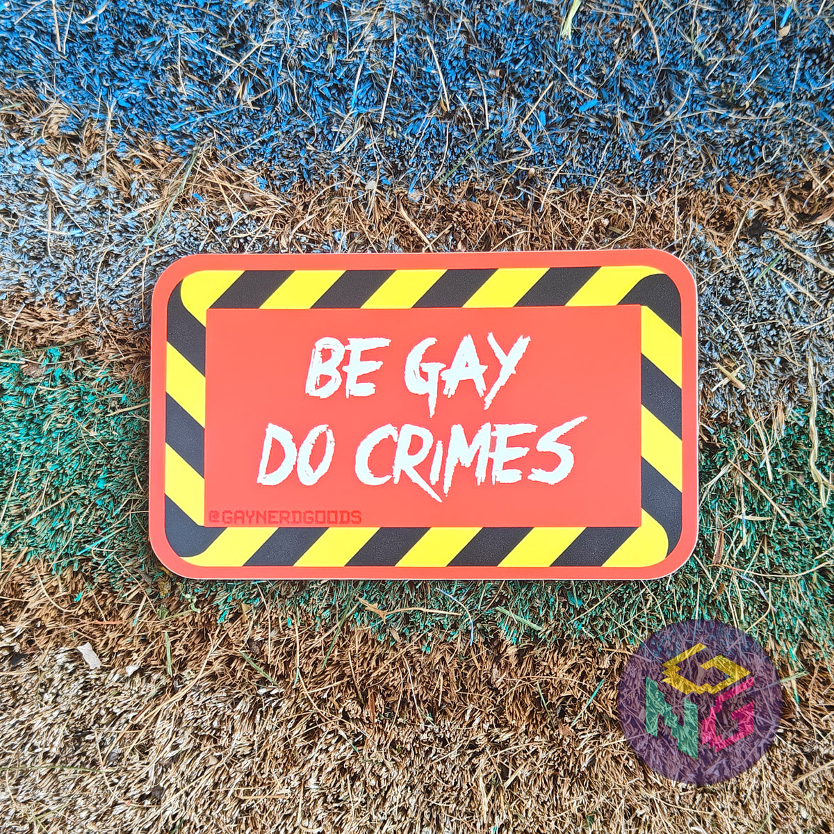Rectangular red sticker featuring white text reading "BE GAY DO CRIMES" surrounded by black and yellow striped border. The sticker is lying flat on a rough rainbow background