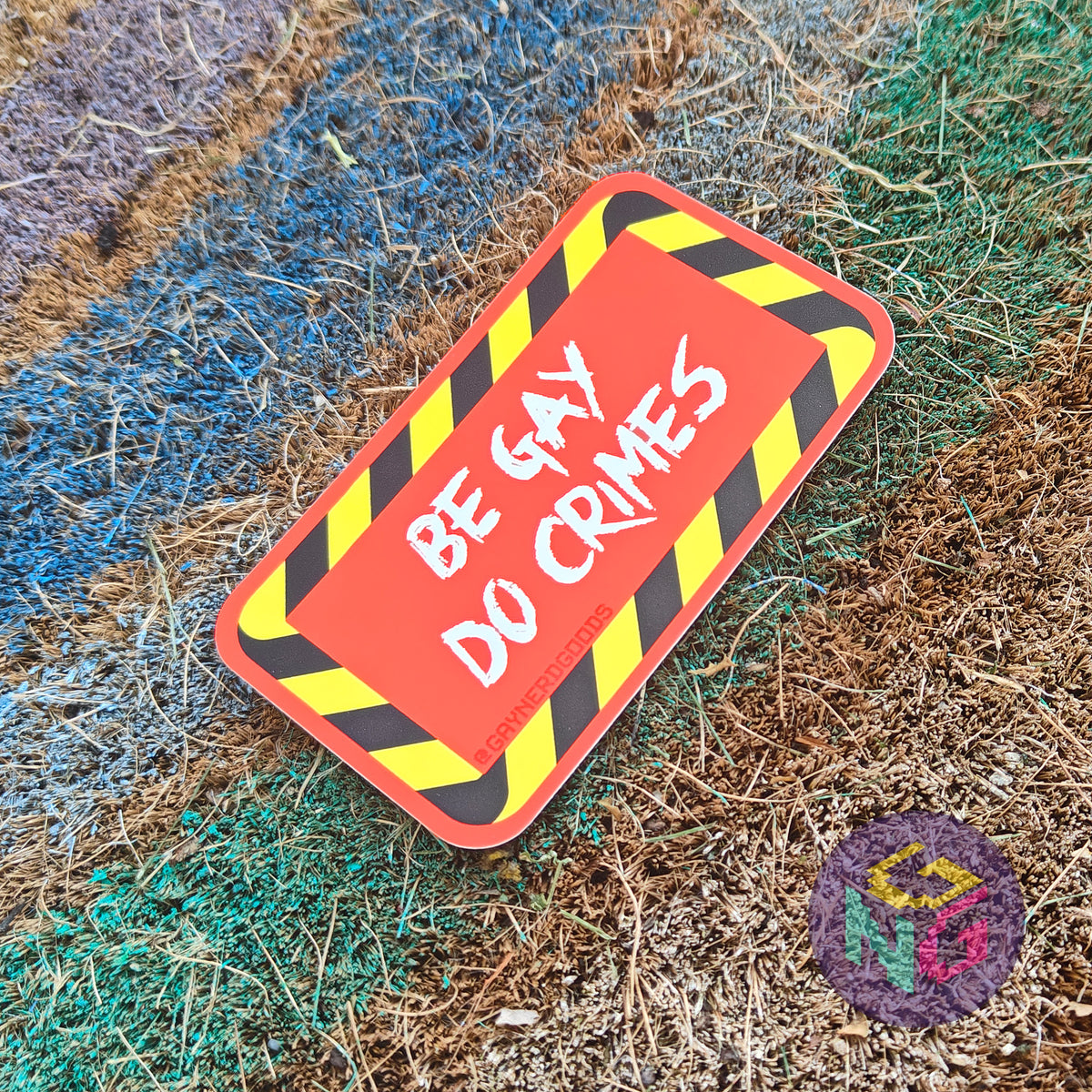 Rectangular red sticker featuring white text reading "BE GAY DO CRIMES" surrounded by black and yellow striped border. The sticker is lying at an angle on a rough rainbow background