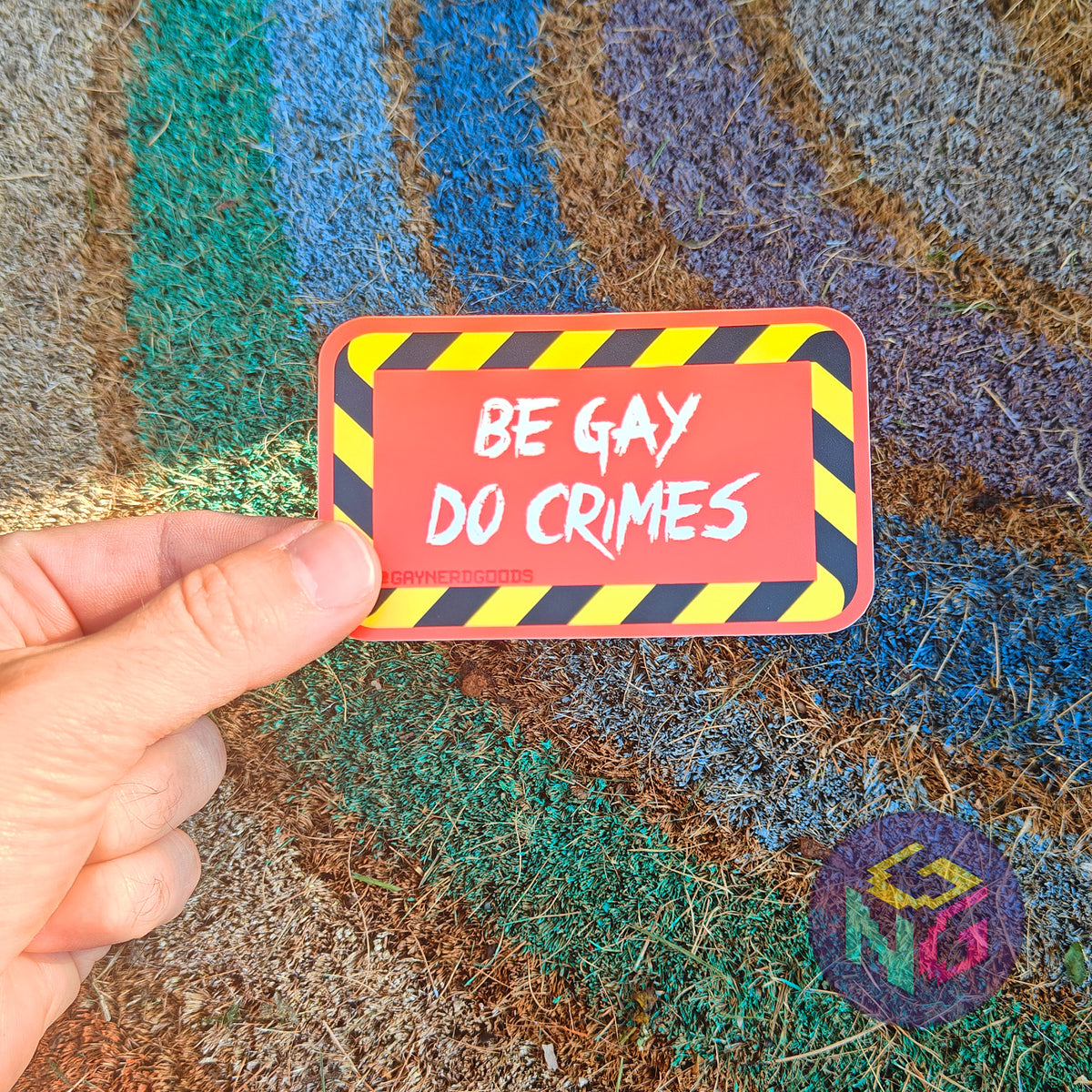 Rectangular red sticker featuring white text reading "BE GAY DO CRIMES" surrounded by black and yellow striped border. The sticker is being held between thumb and forefinger in front of a rough rainbow background