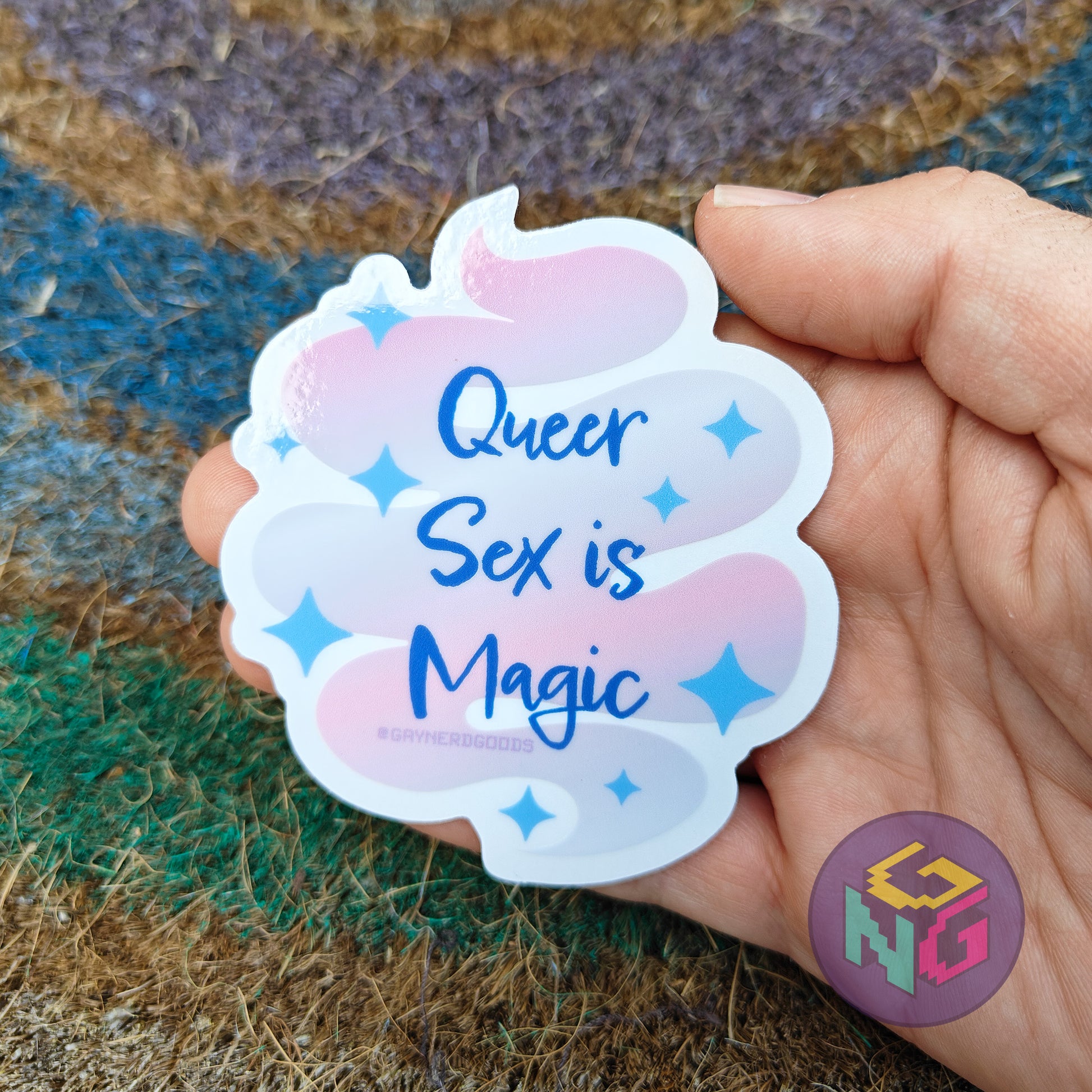 Hand holding a sticker with 'Queer Sex is Magic' text on a textured surface
