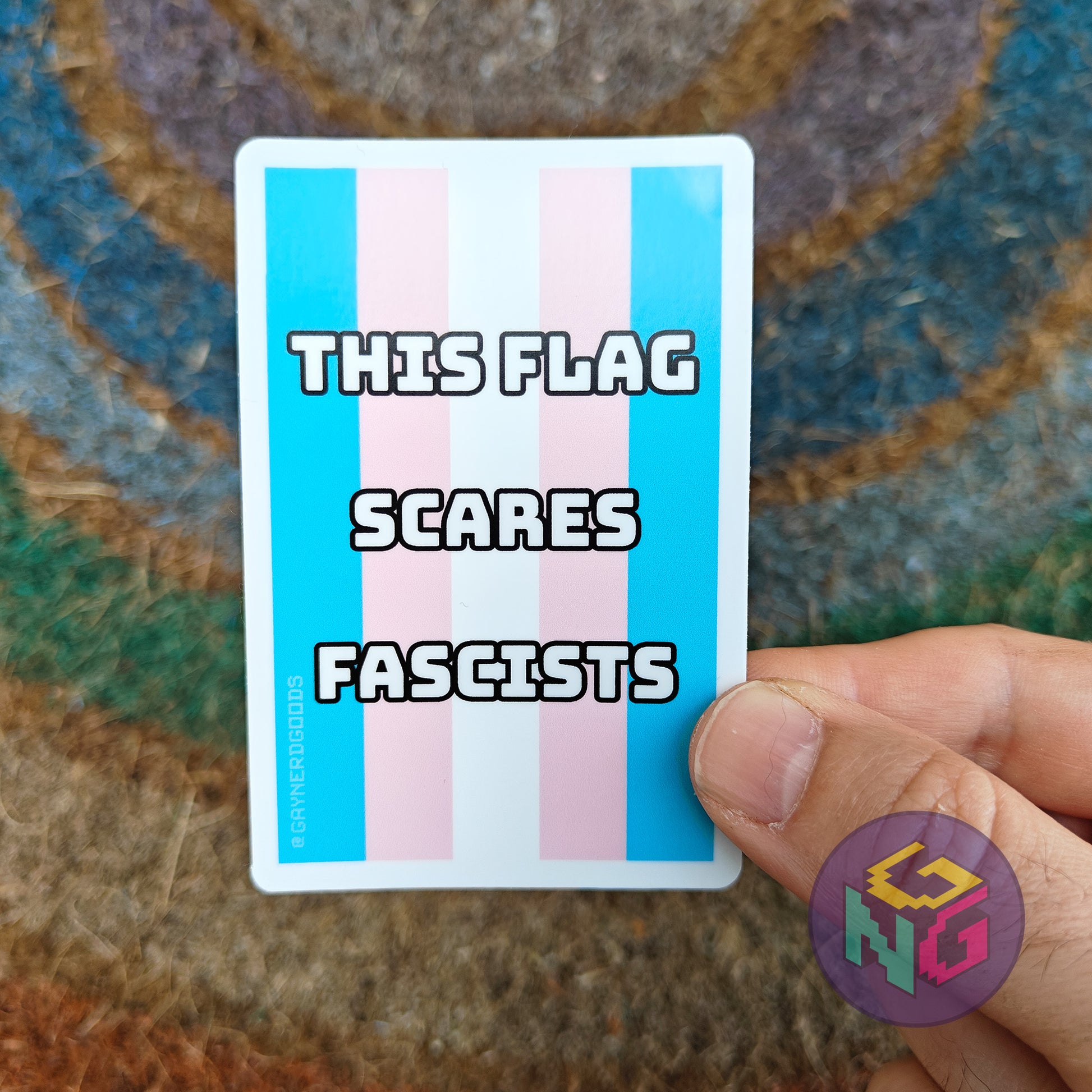 Trans pride flag waterproof sticker with text 'THIS FLAG SCARES FASCISTS' held by a hand on a textured surface.