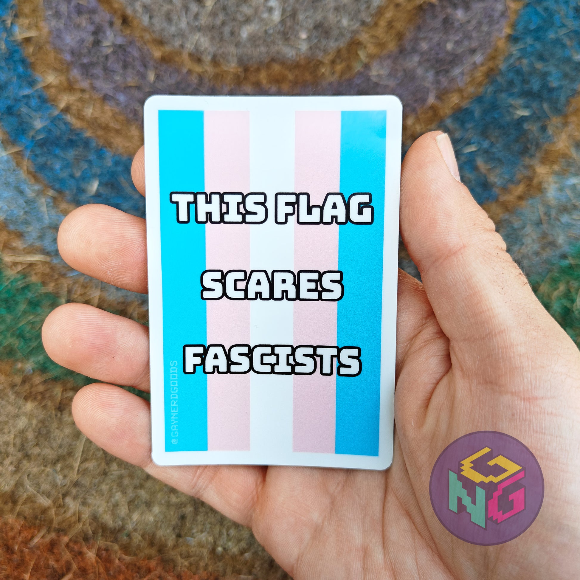 Hand holding a dishwasher safe sticker with a transgender flag and text 'THIS FLAG SCARES FASCISTS' against a textured background.