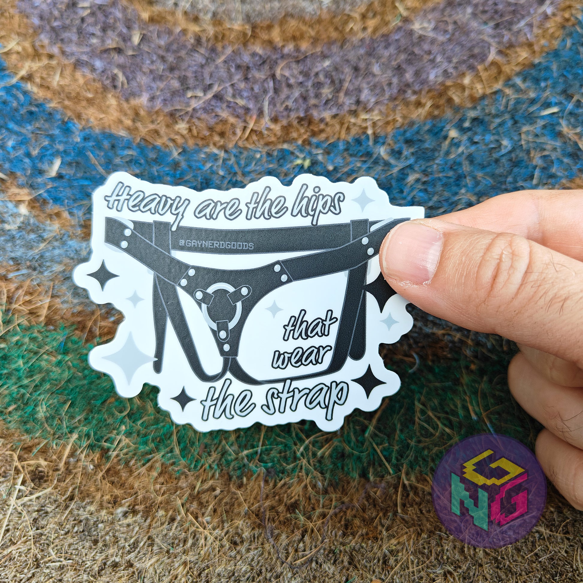 lesbian sex toy harness sticker held in hand surrounded by "heavy are the hips that wear the strap"