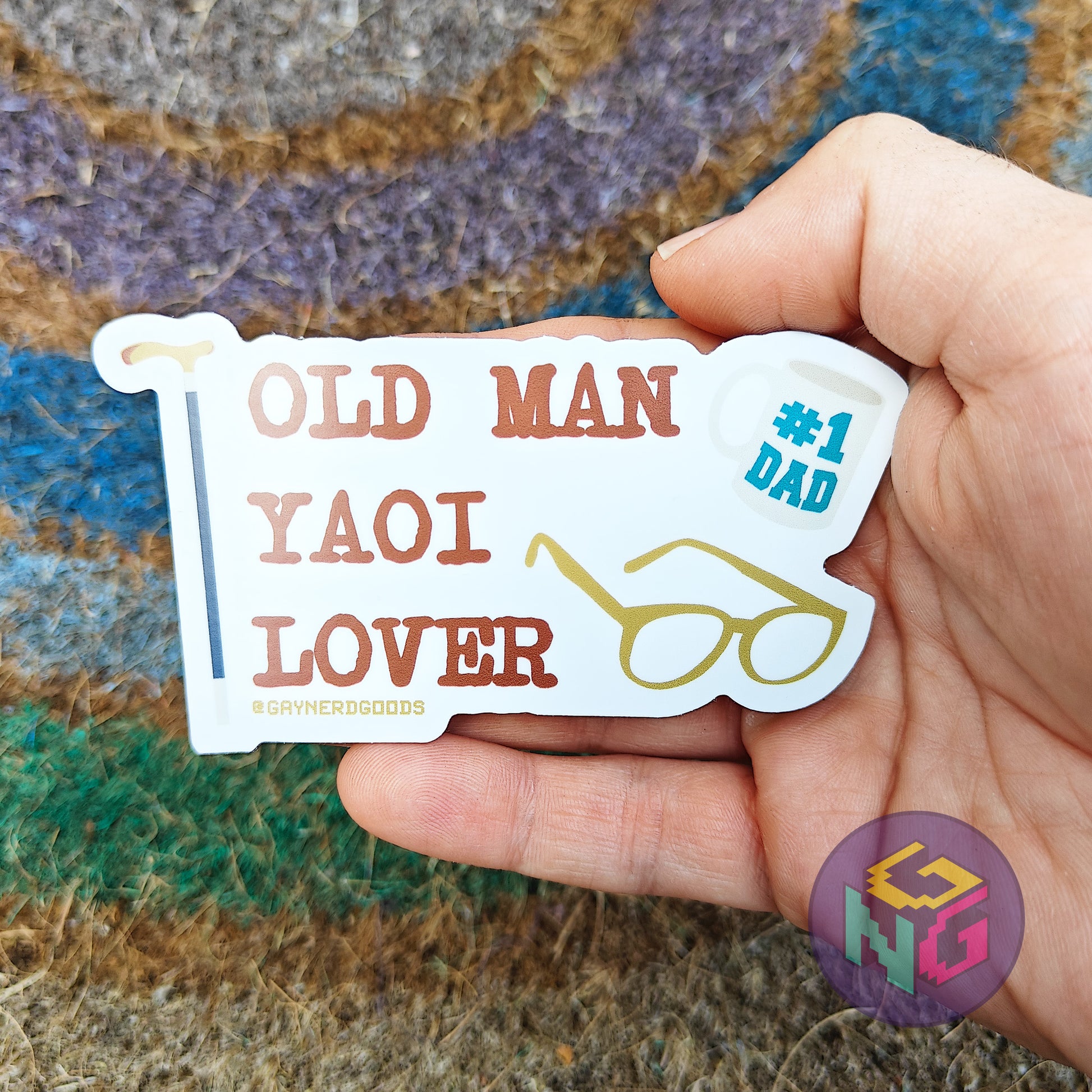 Hand holding a sticker with text 'OLD MAN YAOI LOVER', cane, #1 dad mug, and glasses against a textured background