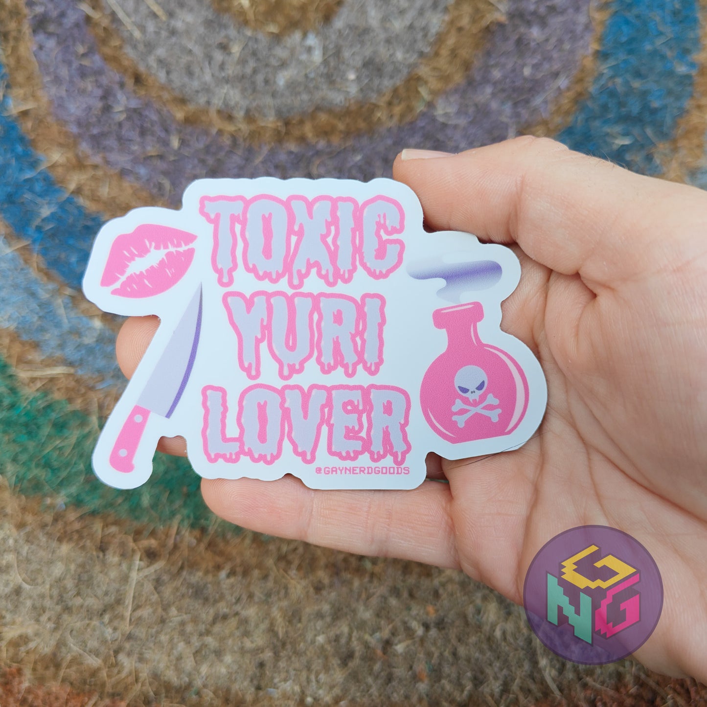 Hand holding a sapphic 'Toxic Yuri Lover' dishwasher safe sticker with a colorful background