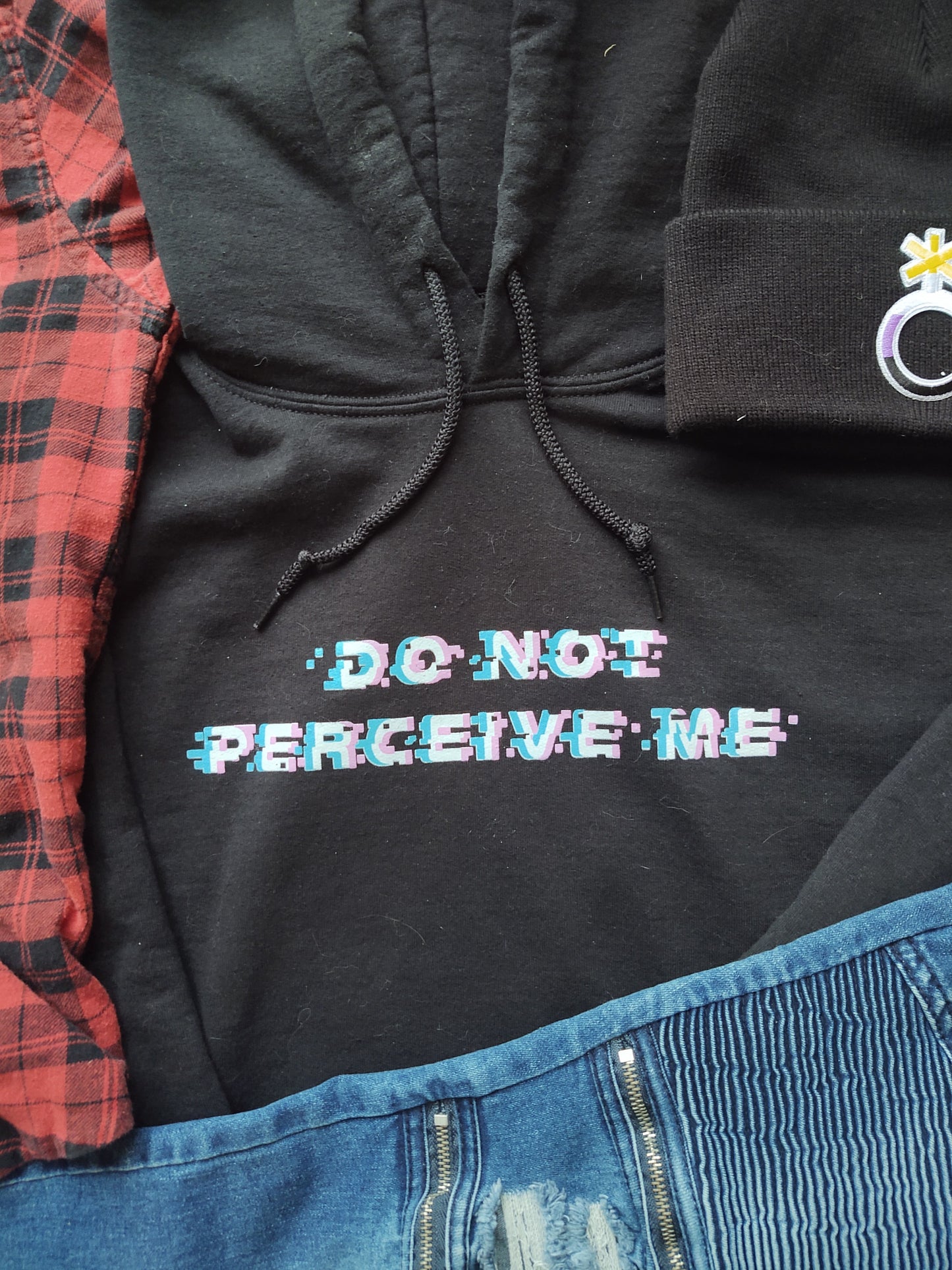 close up of do not perceive me hoodie design surrounded by jeans, flannel, and nonbinary beanie