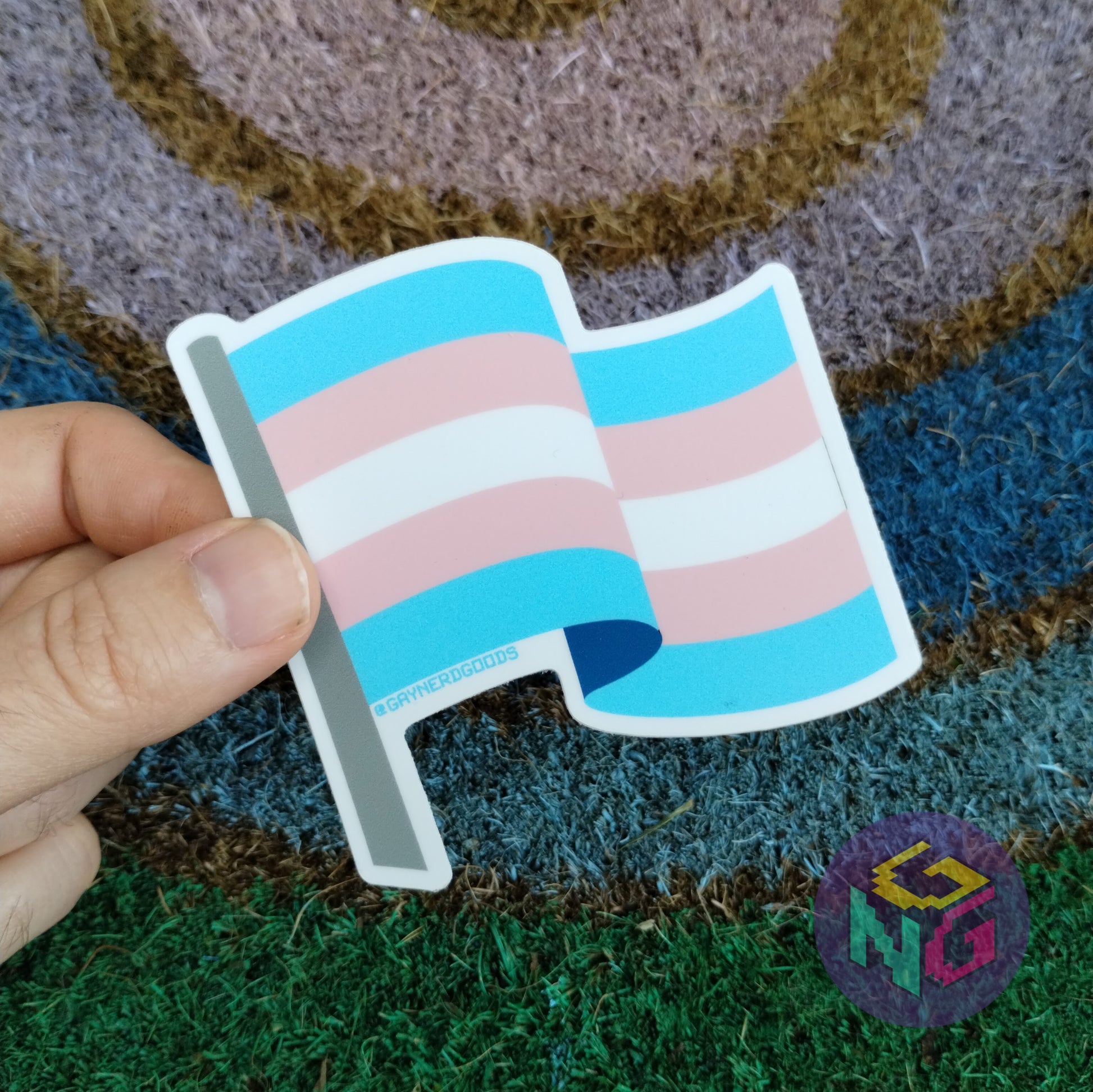 transgender flat sticker held in fingers against rainbow welcome mat