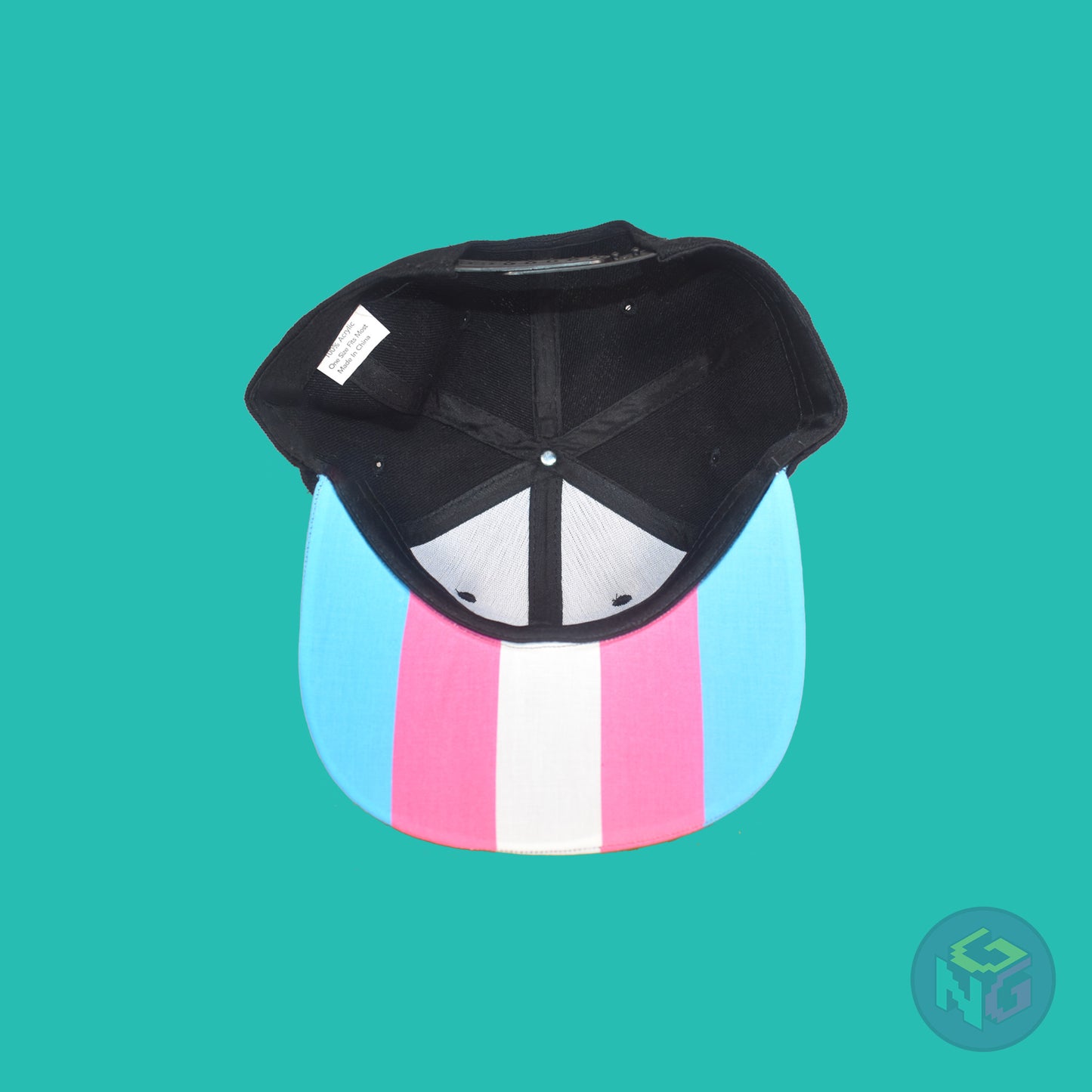 Black flat bill snapback hat. The brim has the transgender pride flag on both sides and the front of the hat has the word “TRANS” in blue, pink, and white letters. Underside view