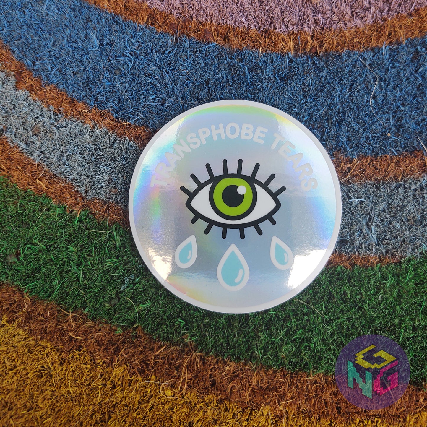rainbow holographic transphobe tears sticker lying flat on rainbow welcome mat