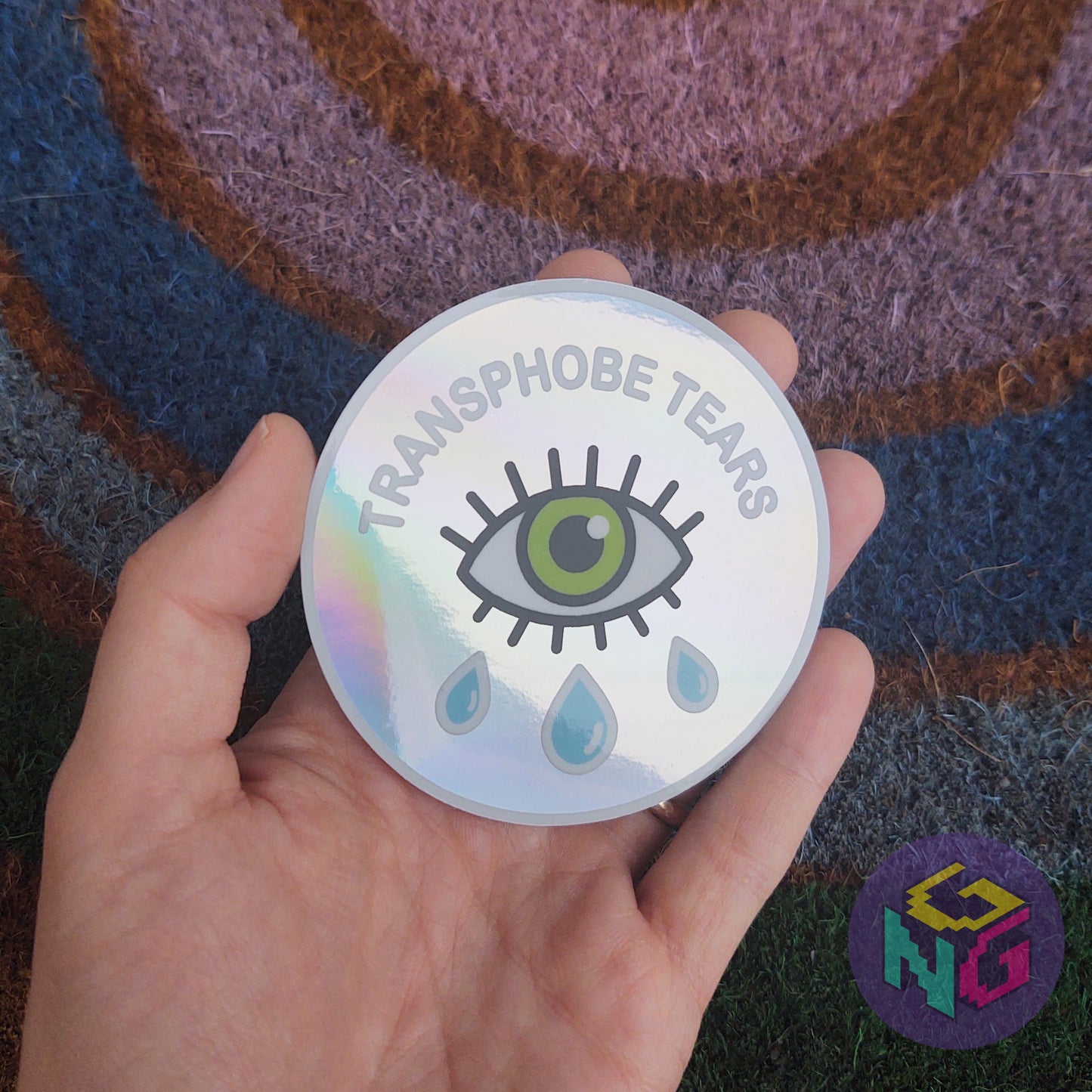 rainbow holographic transphobe tears sticker held in hand in front of rainbow welcome mat