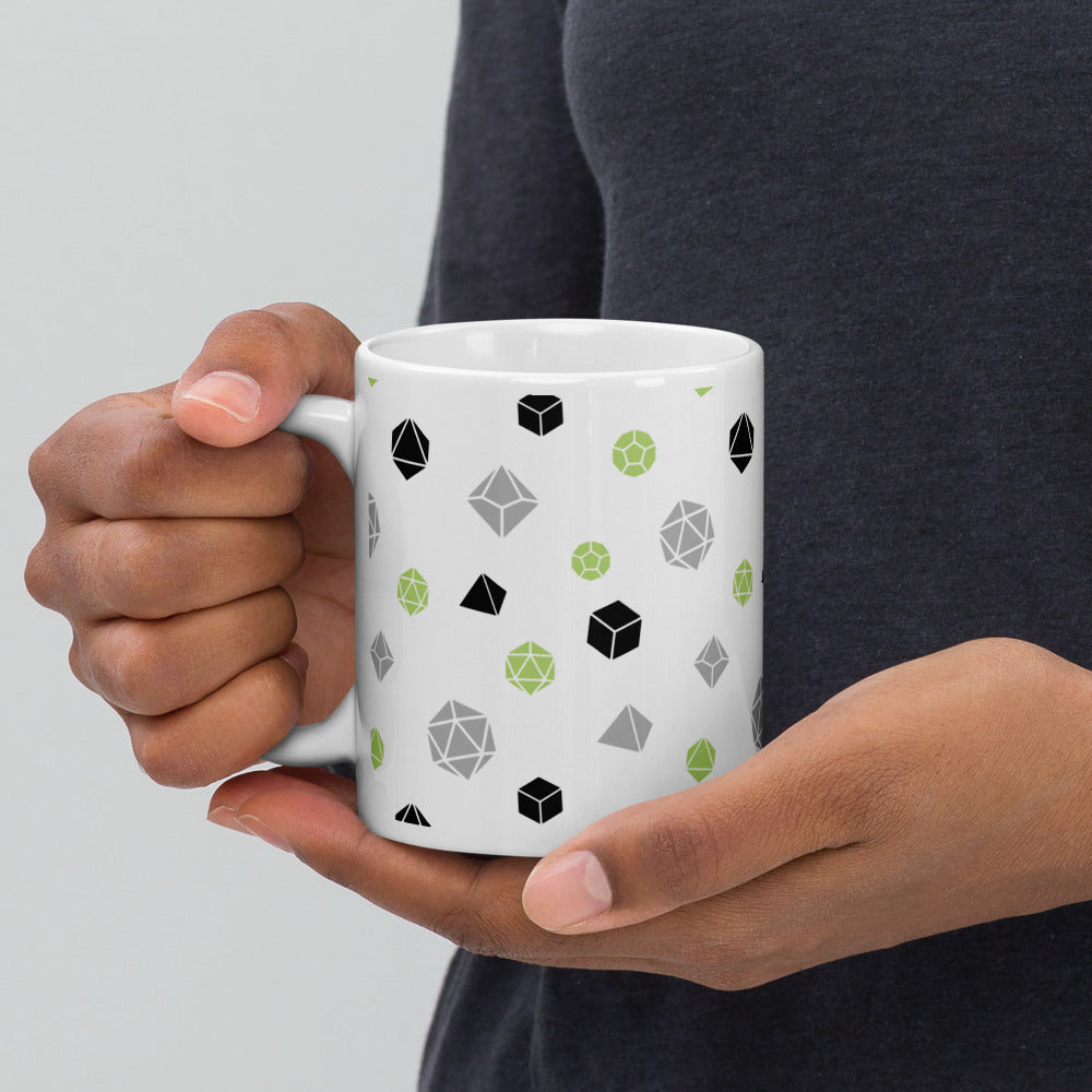 white mug being held by the handle by a dark-skinned set of hands. It has an all-over print of polyhedral d&d dice in the agender colors of green, grey, and black